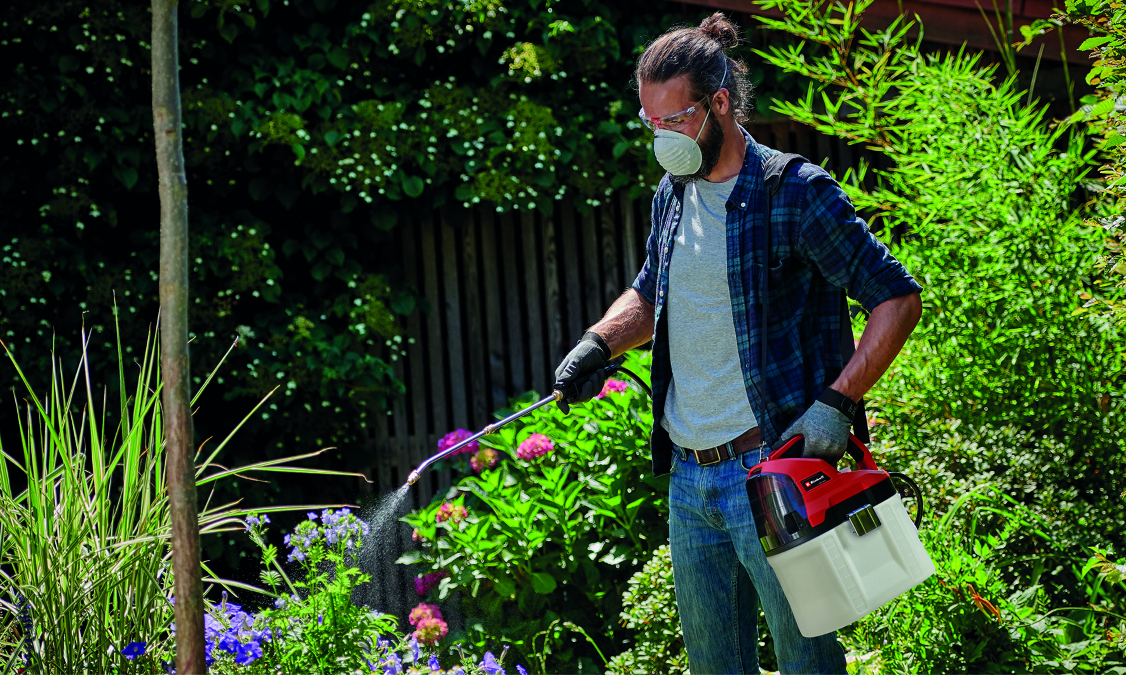 Mann besprüht Pflanzen im Garten. Er trägt eine Maske und Handschuhe und hält ein Sprühgerät und einen Tank.