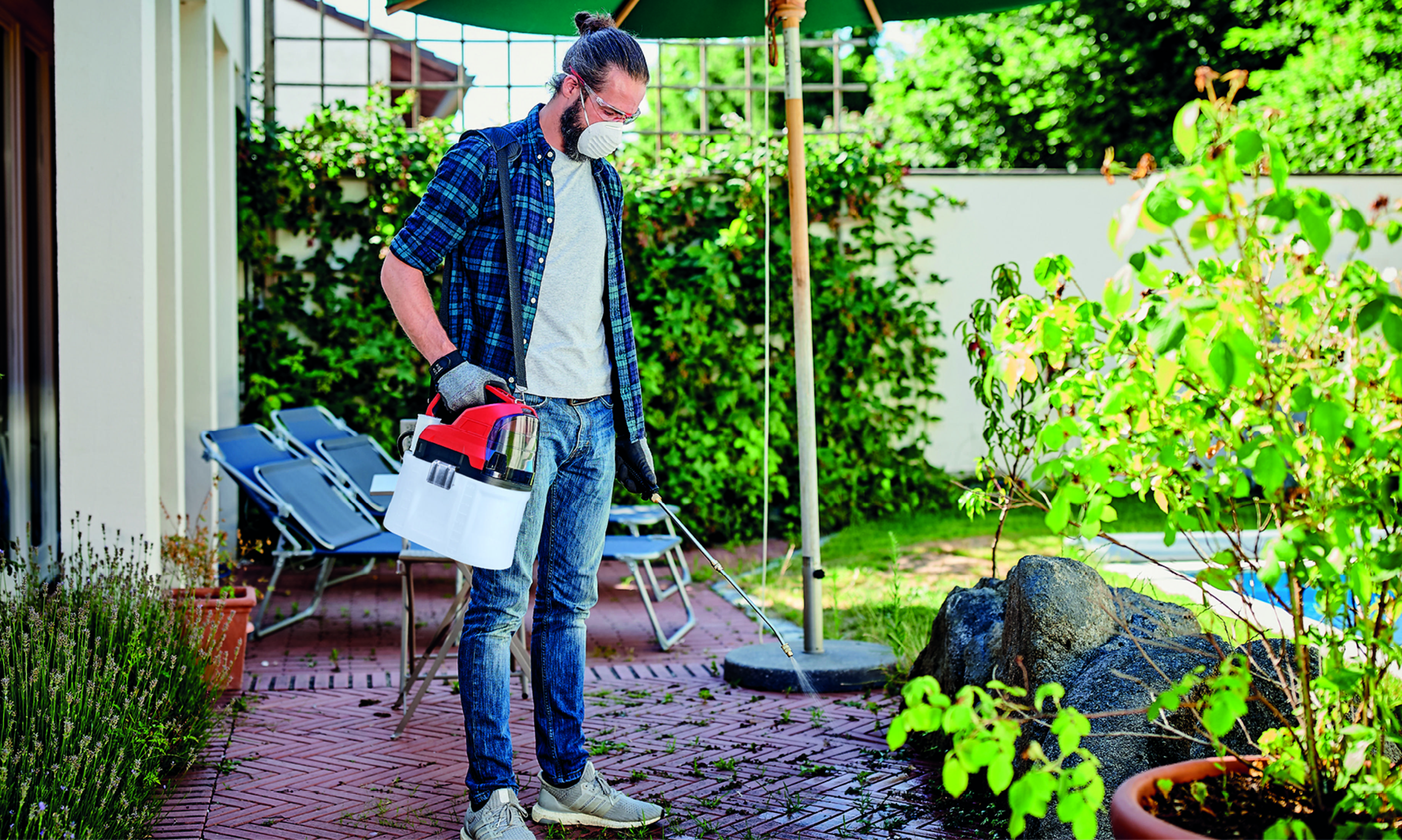 Mann besprüht Pflanzen im Garten. Er trägt eine Maske und hält ein Sprühgerät. Grüne Pflanzen und eine Backsteinterrasse sind zu sehen.