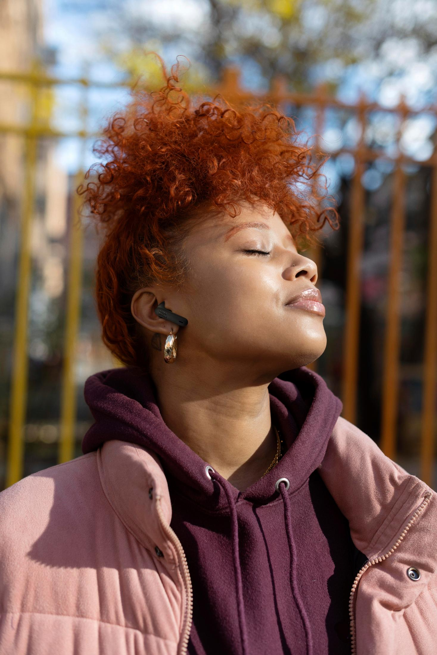 Kobieta z rudymi kręconymi włosami, zamkniętymi oczami, w różowej kamizelce i fioletowej bluzie z kapturem. Ma złoty kolczyk i jest na zewnątrz.