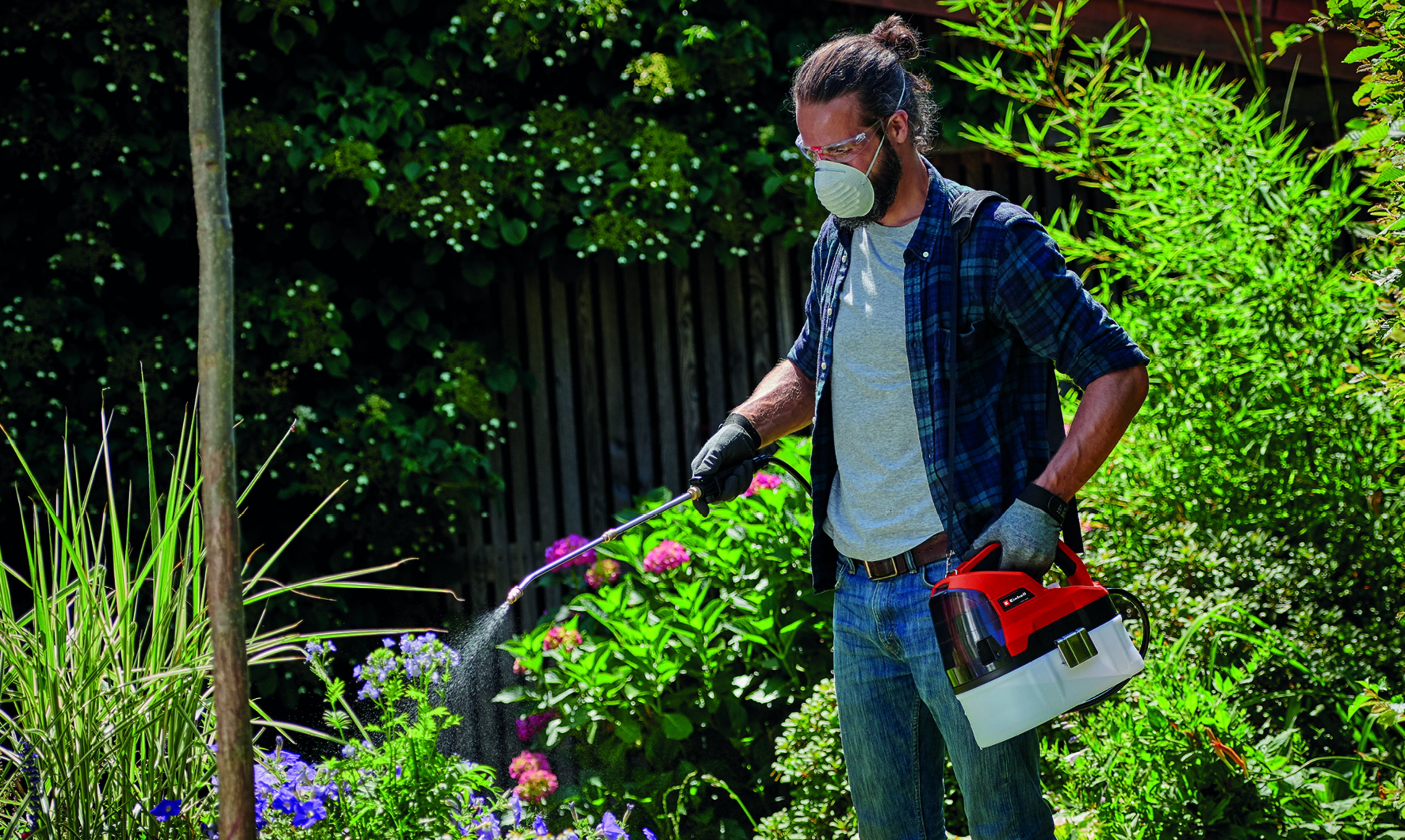 Ein Mann im Garten besprüht Pflanzen mit einem rot-weißen Sprühgerät, trägt Maske, Brille und Handschuhe. Grünes Laub im Hintergrund.