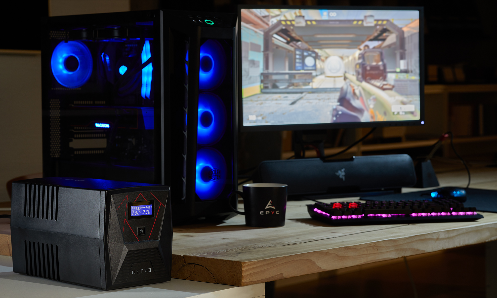 Un'installazione di gioco con PC, monitor che mostra un gioco e periferiche. Un UPS è in primo piano. Le luci blu illuminano il PC.