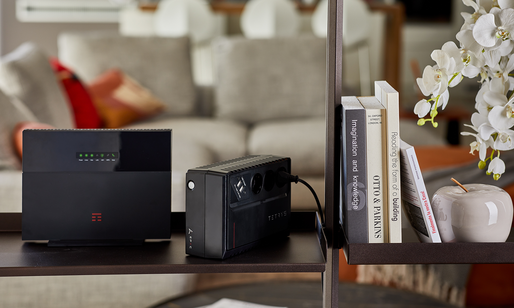 Un dispositivo elettronico nero con luci, una ciabatta nera, libri, fiori bianchi e una mela grigia su una mensola.