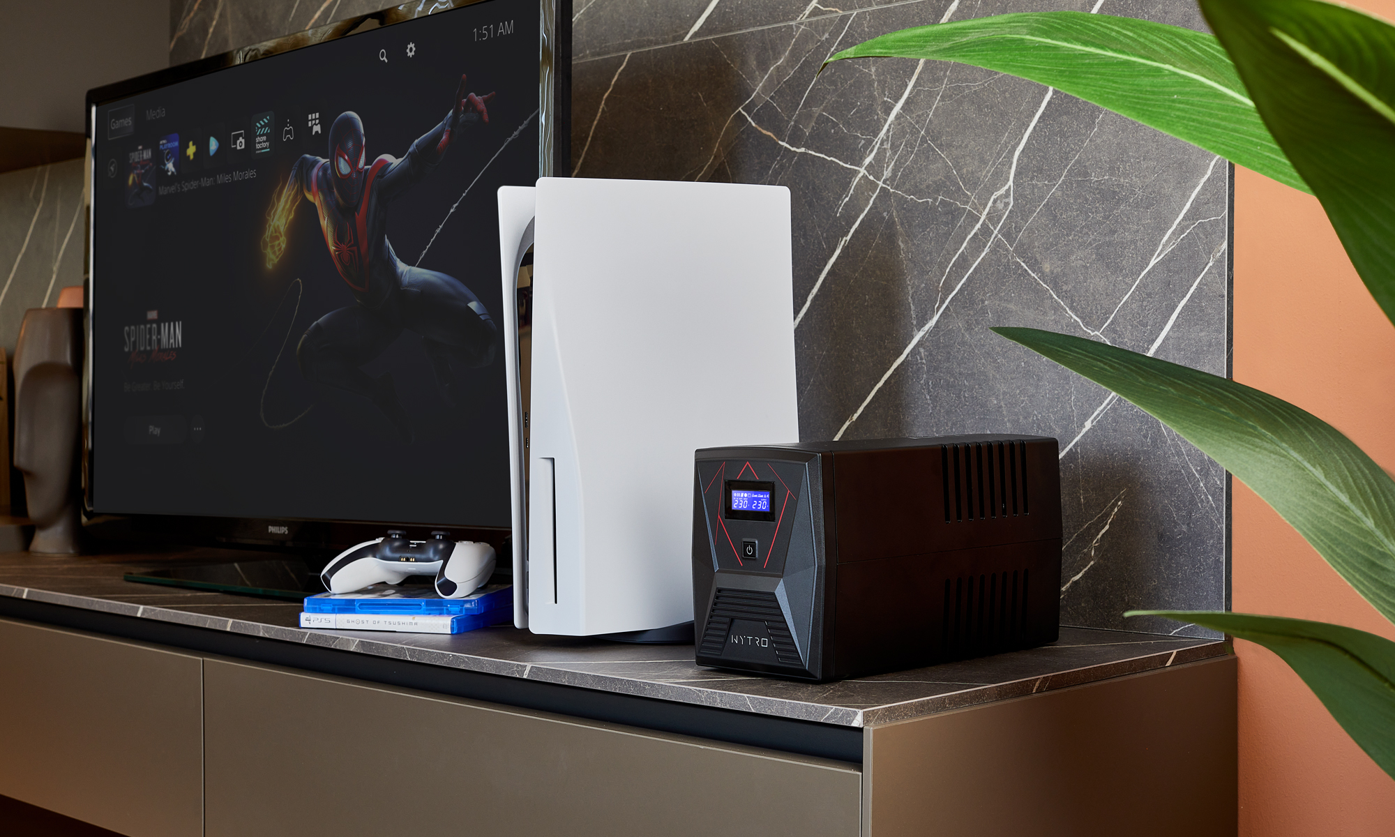Una TV mostra il gioco di Spider-Man, con una PS5, un controller e un UPS nero. Una pianta è a destra. Un muro con motivo marmoreo sullo sfondo.