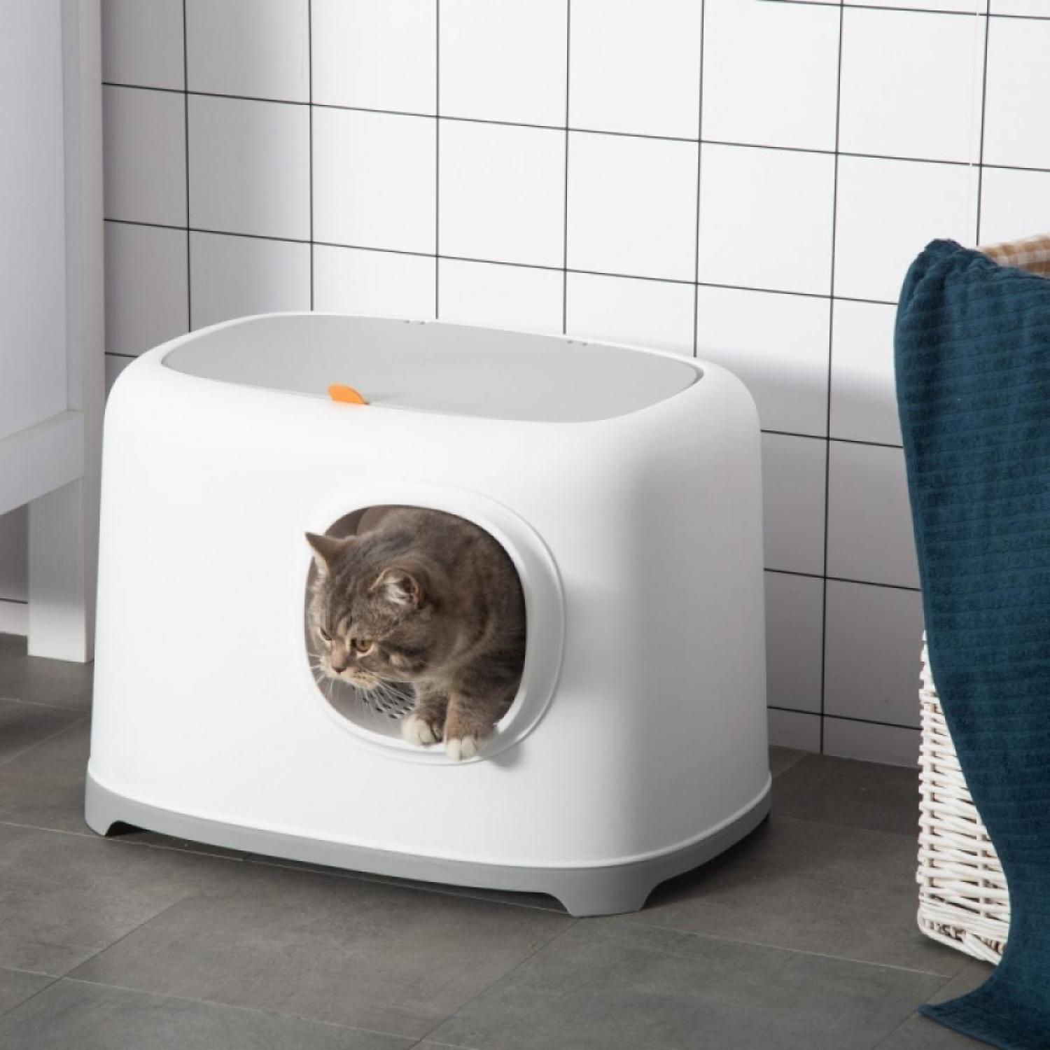 Eine graue Katze verlässt eine weiße Katzentoilette. Die Box befindet sich in einem Badezimmer mit weißen Fliesen. Rechts sind ein Korb und ein Handtuch.