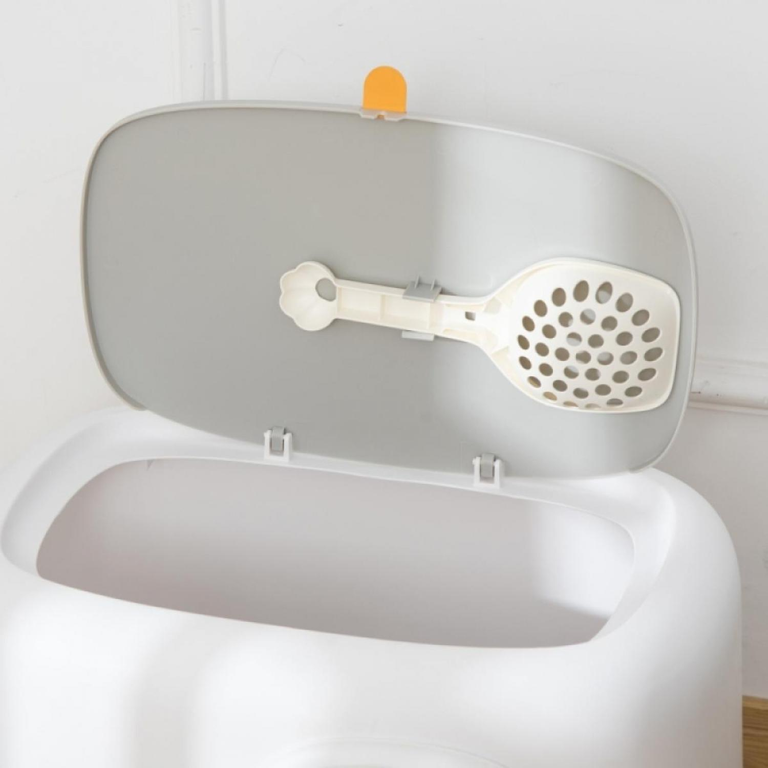 Eine weiße und graue Katzentoilette mit offenem Deckel, einer Schaufel und einem orangefarbenen Riegel. Die Schaufel ist am Deckel befestigt.