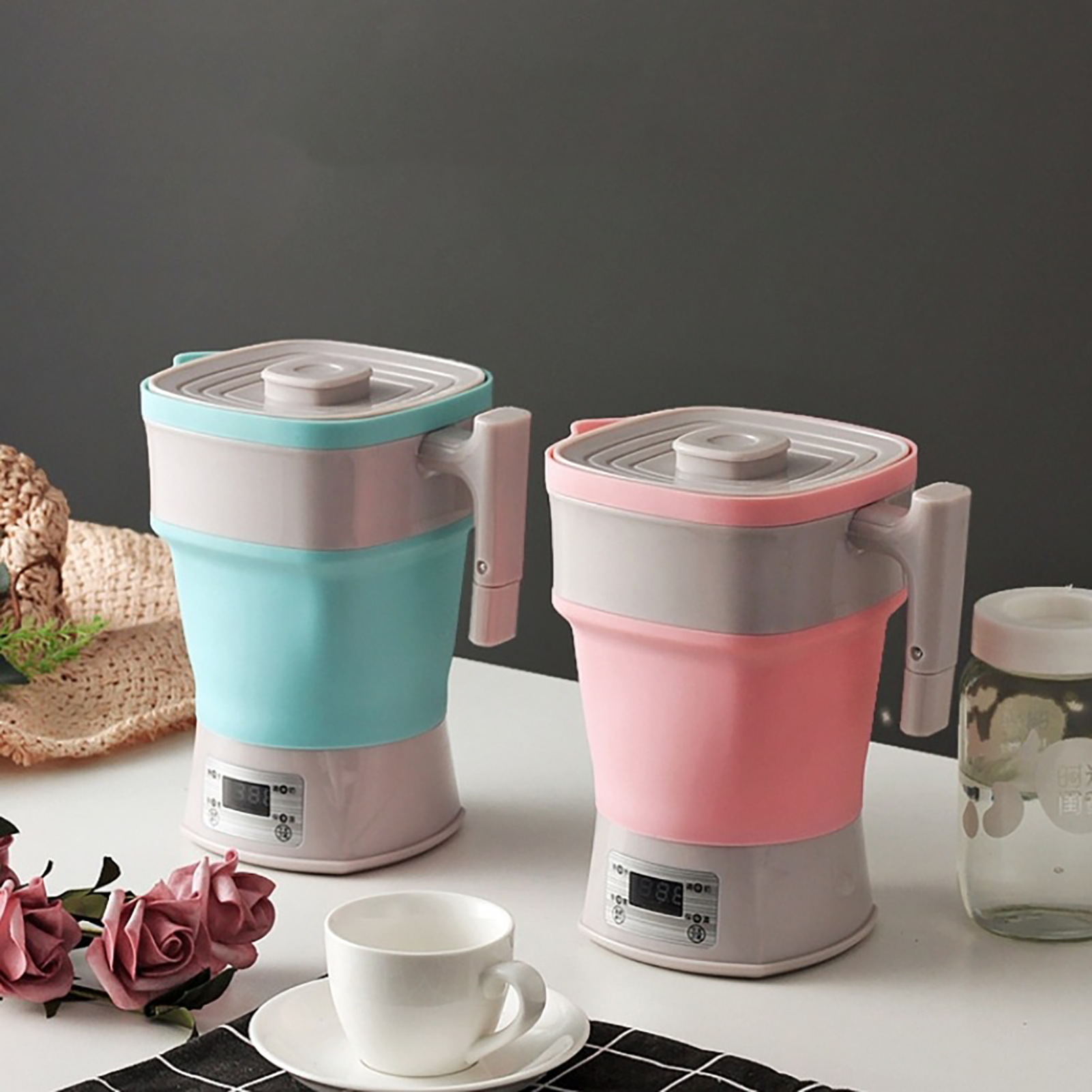 Zwei bunte elektrische Wasserkocher, blau und rosa, stehen auf einem weißen Tisch. Eine Tasse, Blumen und ein Glas sind ebenfalls vorhanden.