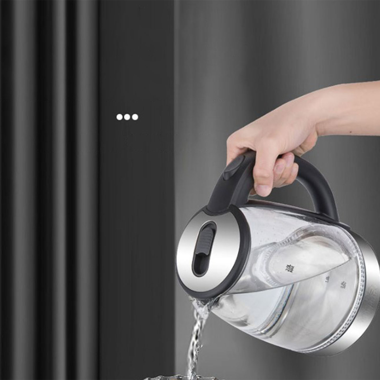 Eine Hand gießt Wasser aus einem transparenten Wasserkocher mit schwarzem Griff. Der Hintergrund hat ein verschwommenes, dunkles, vertikales Muster.