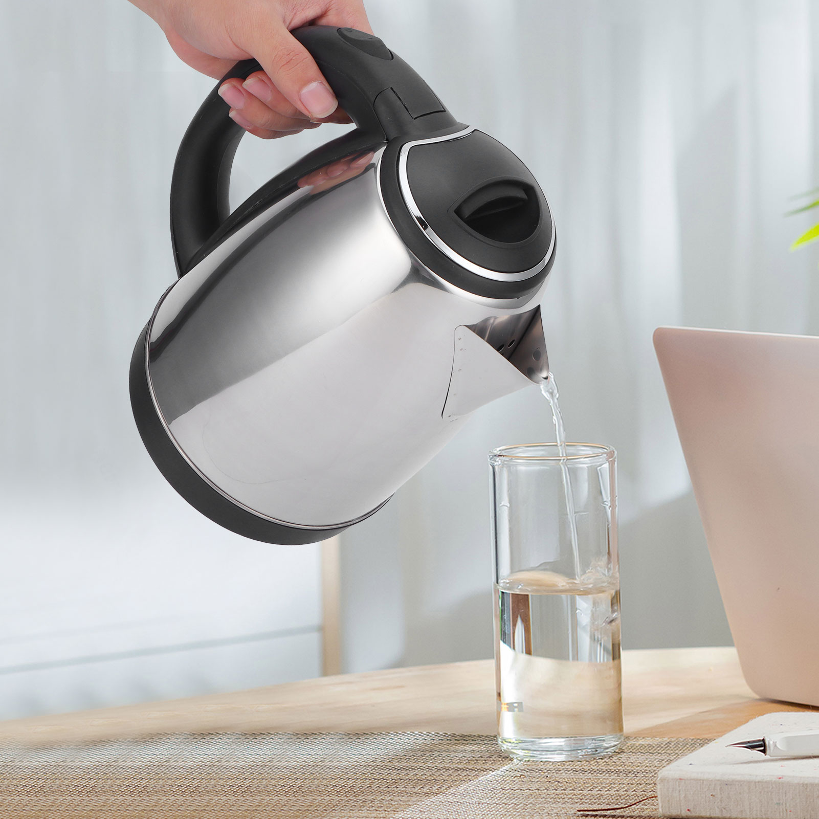 Eine Hand gießt Wasser aus einem Edelstahlkessel in ein Glas. Auf der rechten Seite ist ein beiger Laptop zu sehen.