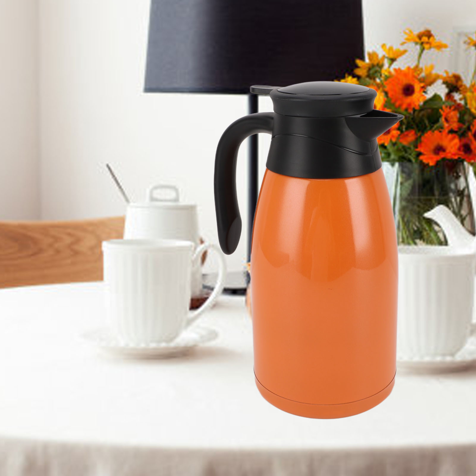 Eine orangefarbene und schwarze Thermoskanne auf einem weißen Tisch mit weißen Tassen und einer Blumenvase im Hintergrund.