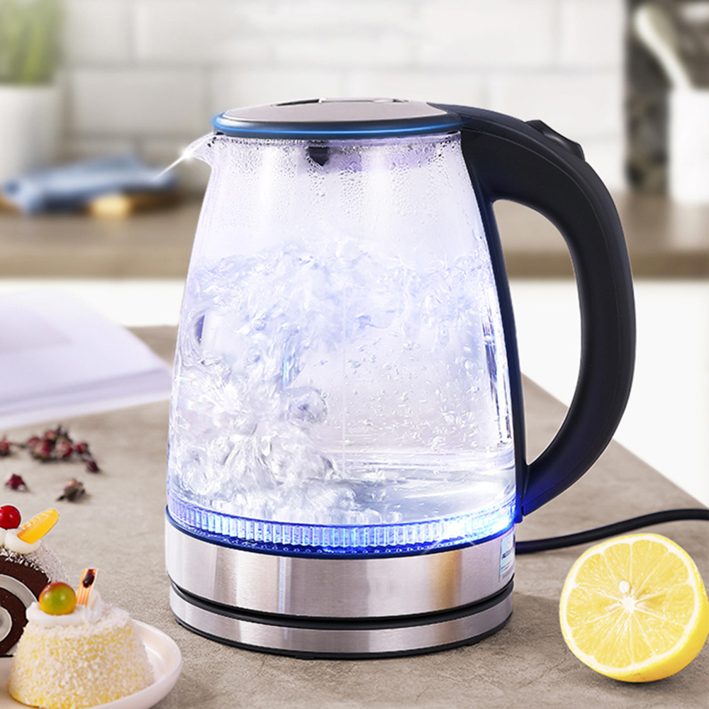 Ein elektrischer Glaskessel mit kochendem Wasser. Eine Zitronenhälfte und Gebäck auf dem Tisch. Schwarzer Griff und ein blau leuchtender Sockel.