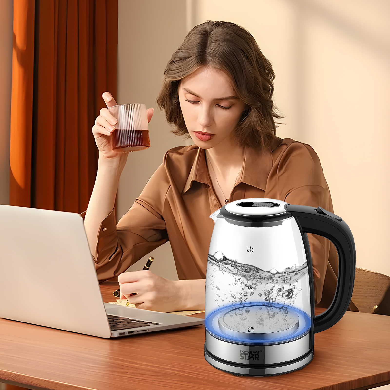 Frau mit einem Glas Getränk, arbeitet an einem Laptop. Auf dem Tisch steht ein Glaskessel mit kochendem Wasser.