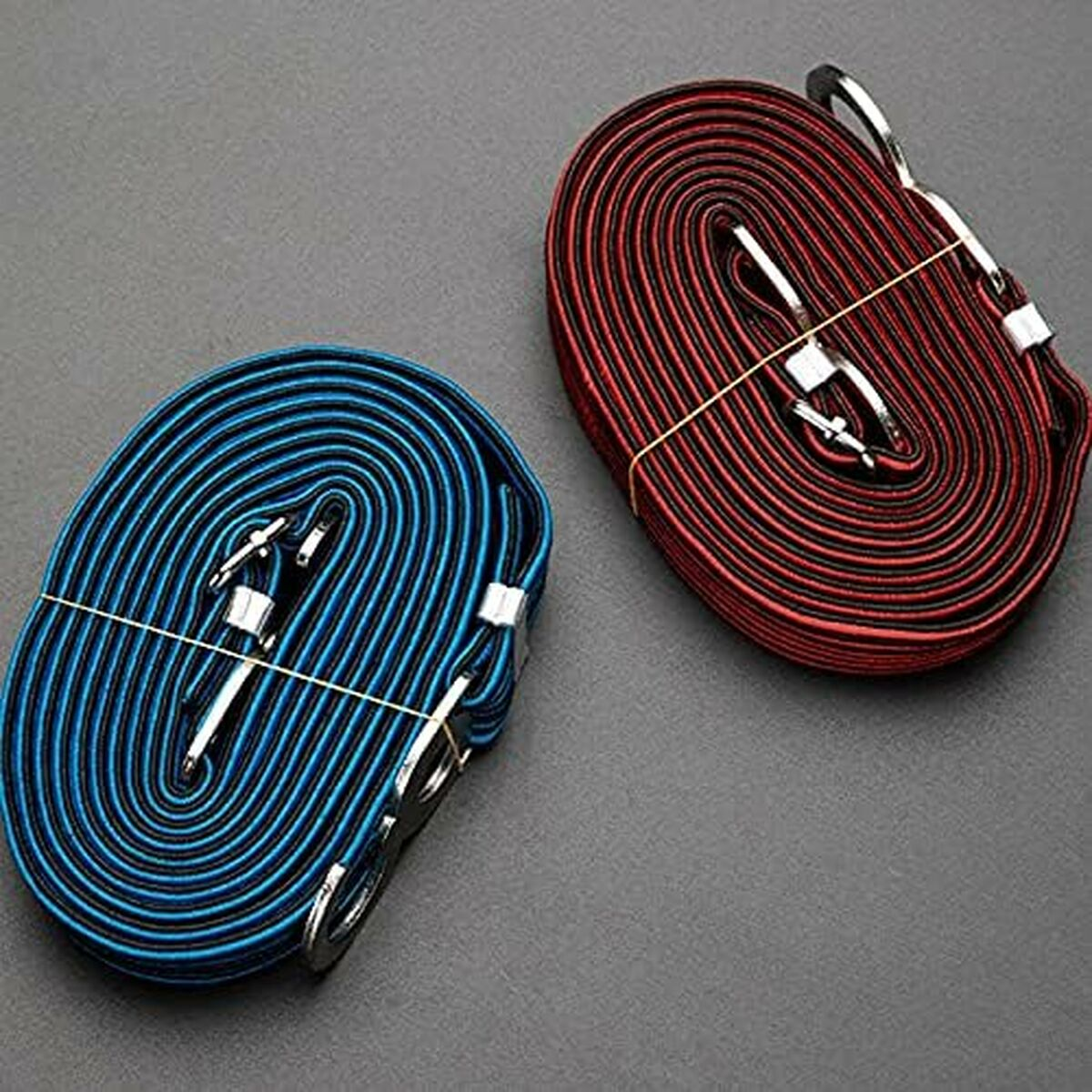 Zwei aufgerollte Bungee-Seile, eines blau und eines rot, mit Metallhaken, liegen auf einer grauen Oberfläche. Jedes ist mit einem Gummiband gebunden.