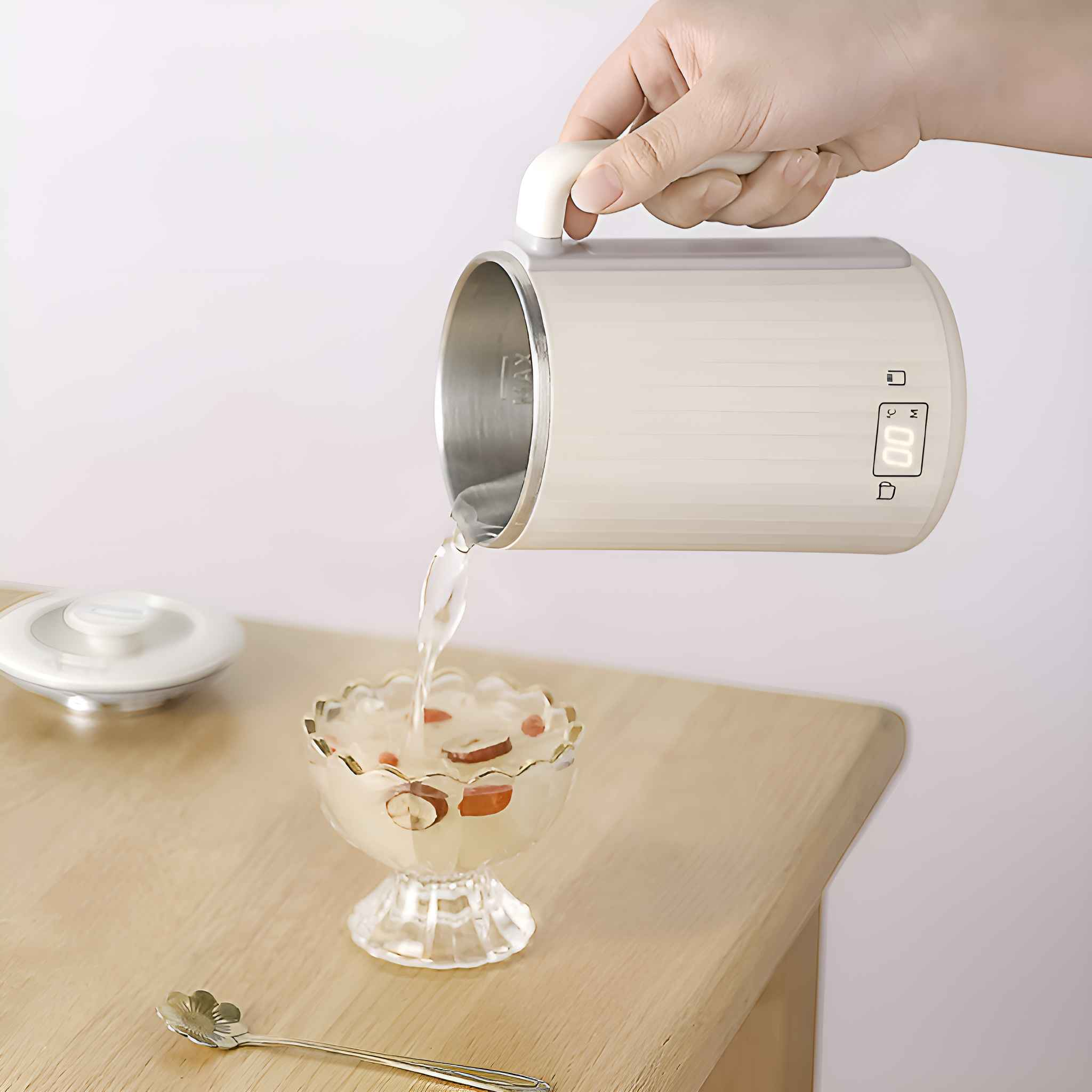 Eine Hand gießt Wasser aus einem weißen Wasserkocher in eine Glasschale mit Früchten. Ein Löffel liegt daneben.