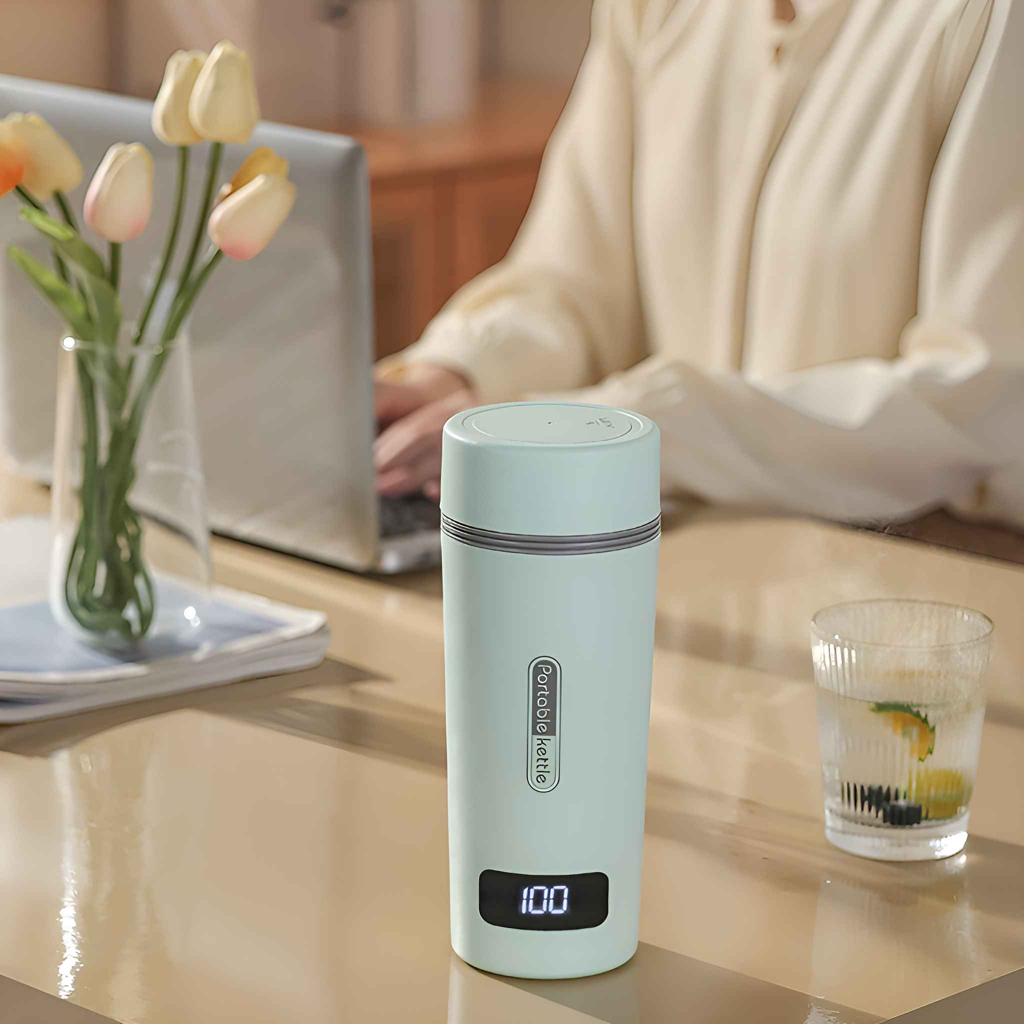 Ein tragbarer türkisfarbener Wasserkocher, ein Glas Wasser und Tulpen auf einem Tisch. Im Hintergrund arbeitet eine Person an einem Laptop.