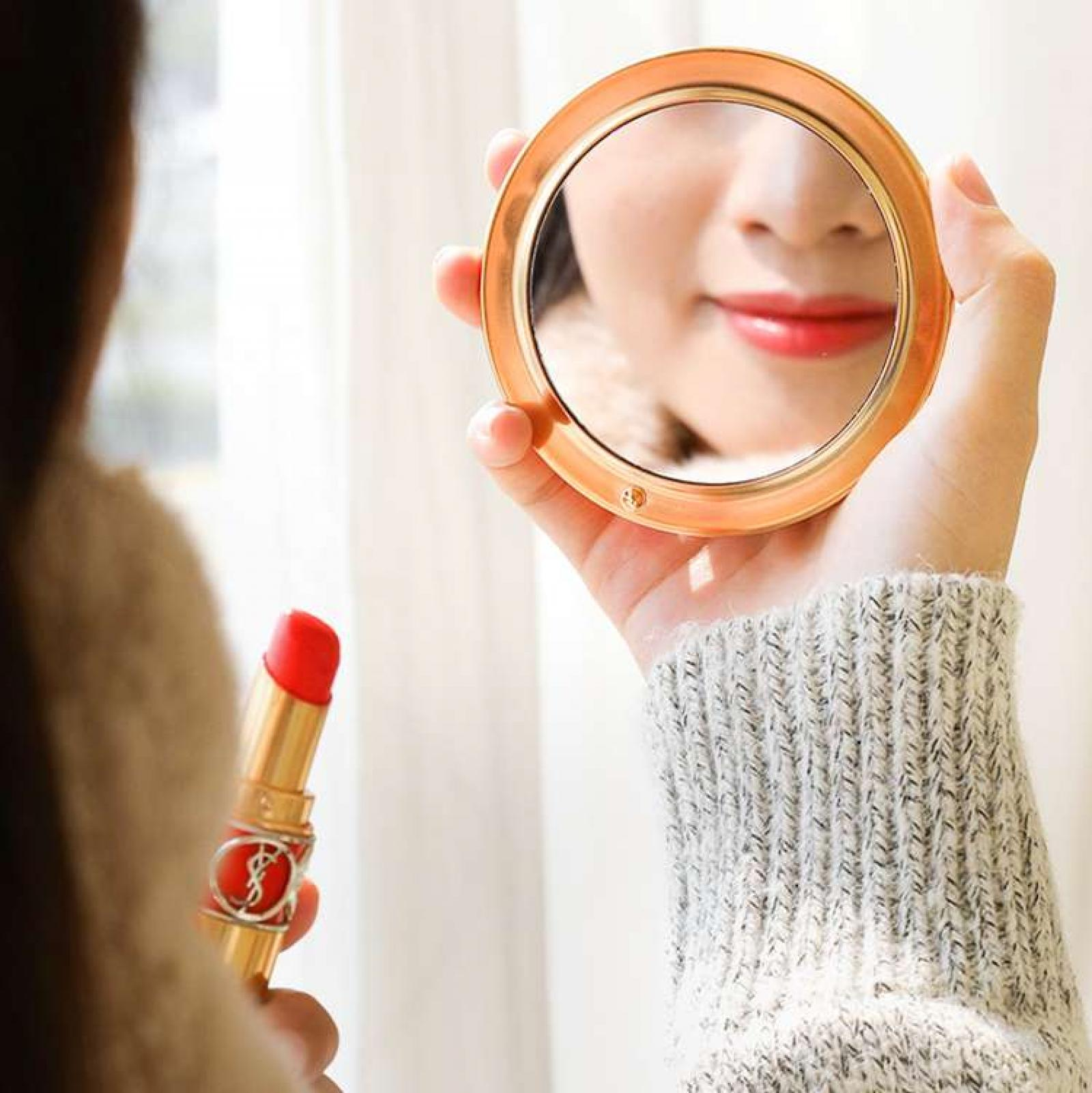 Eine Person hält einen Spiegel, der roten Lippenstift auf Lippen reflektiert. Ein Lippenstift ist in der anderen Hand. Ein grauer Pullover ist sichtbar.