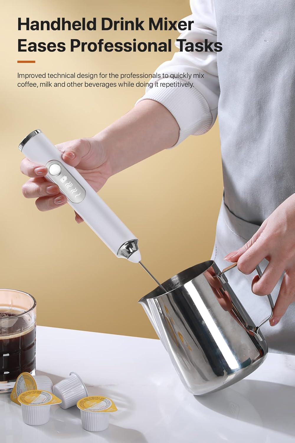 Eine Person verwendet einen weißen Hand-Getränkemixer, um ein Getränk in einem Stahlkrug zu mixen. Kaffeepads und ein Glas Kaffee stehen auf dem Tisch.
