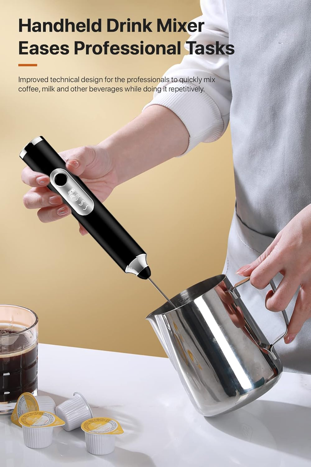 Eine Person verwendet einen schwarzen Handmixer, um Flüssigkeit in einem Metallkrug zu verrühren. Kaffeepads und ein Glas Kaffee sind ebenfalls vorhanden.
