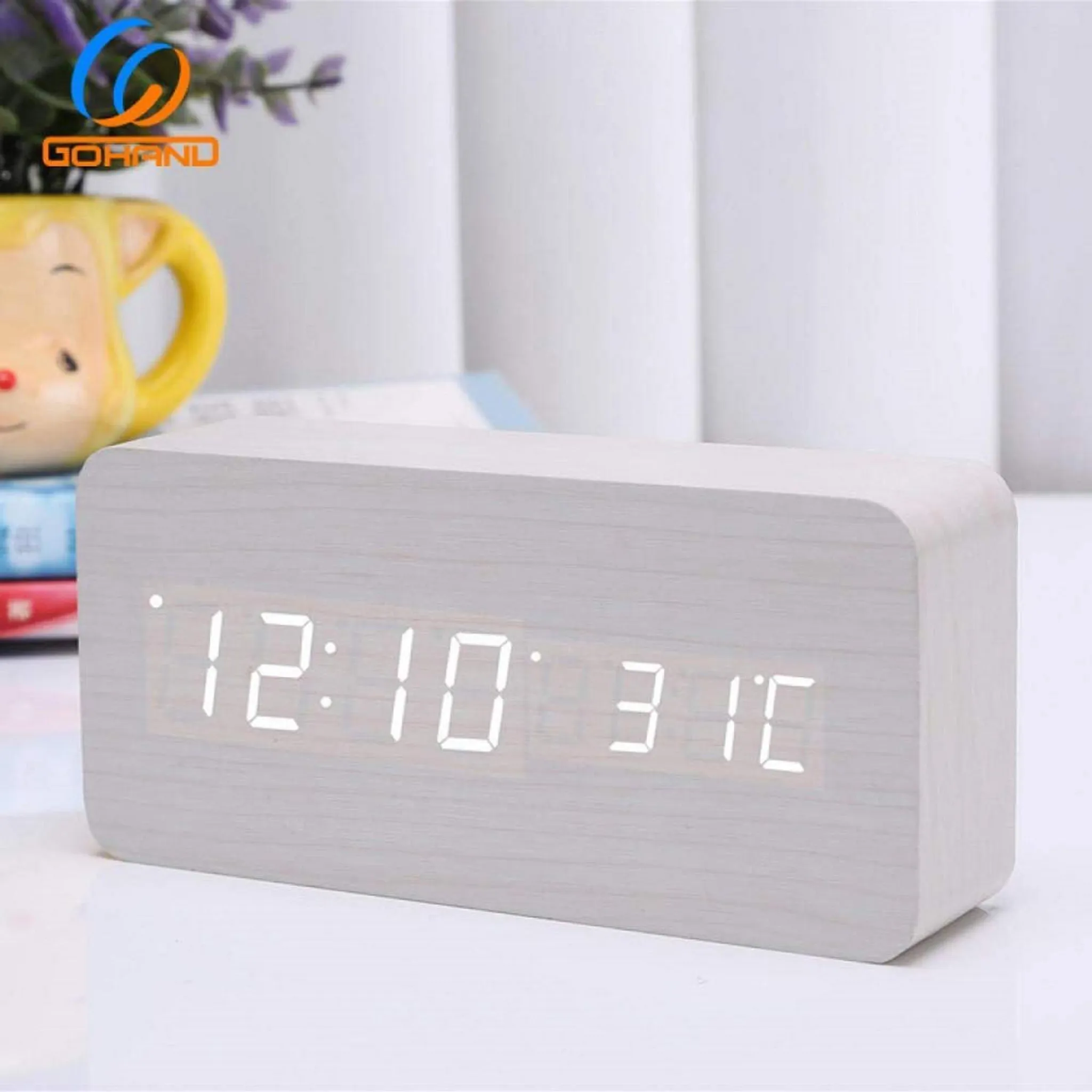 Eine rechteckige Digitaluhr zeigt 12:10 und 31C an. Sie ist hellgrau, aus Holz und steht auf einem Tisch mit verschwommenen Objekten im Hintergrund.