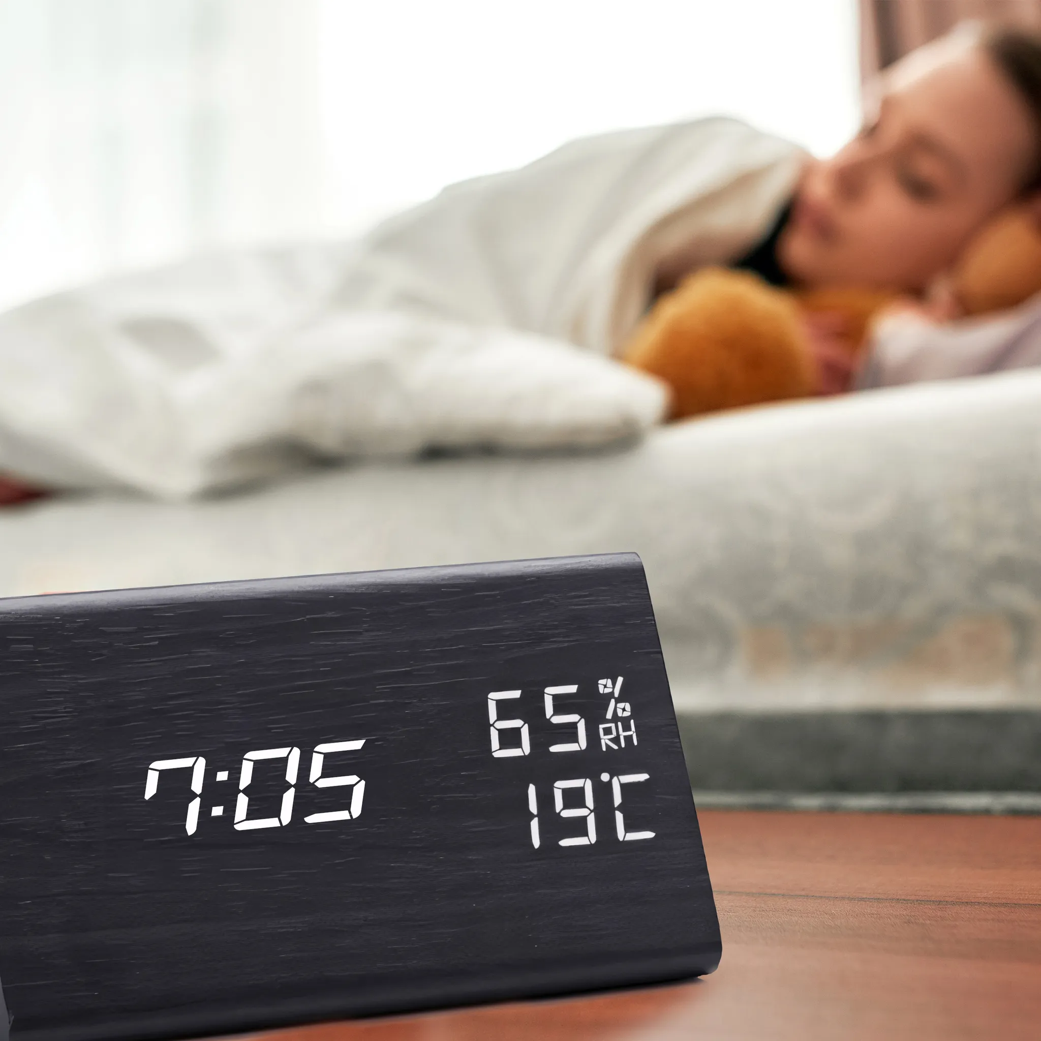 Eine Digitaluhr zeigt 7:05, 65 % Luftfeuchtigkeit und 19 °C an. Eine Person schläft im Bett, mit Kissen und Decke.