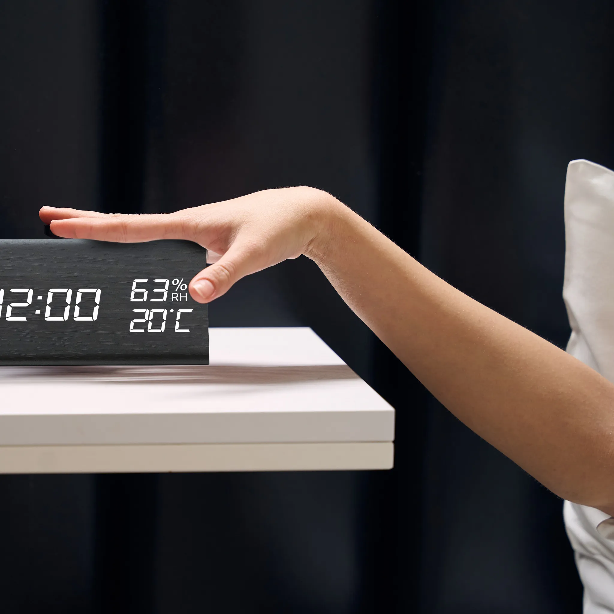 Eine Hand greift nach einer Digitaluhr mit der Anzeige 12:00, Luftfeuchtigkeit und Temperatur. Der Hintergrund ist schwarz, ein weißes Kissen ist sichtbar.