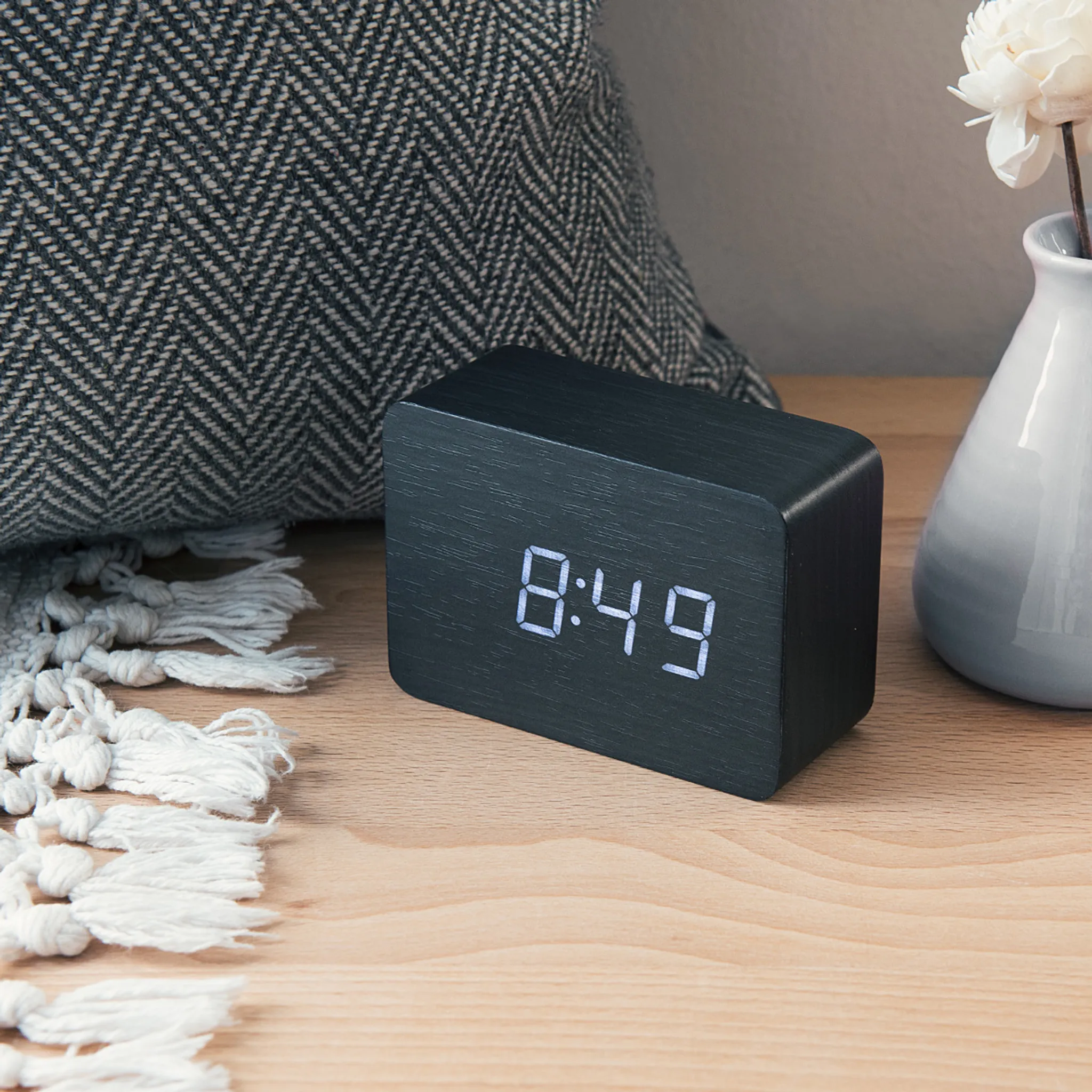 Eine schwarze Digitaluhr zeigt 8:49 an. Ein graues Kissen, weiße Quastendeko und eine Vase mit einer Blume sind im Hintergrund.