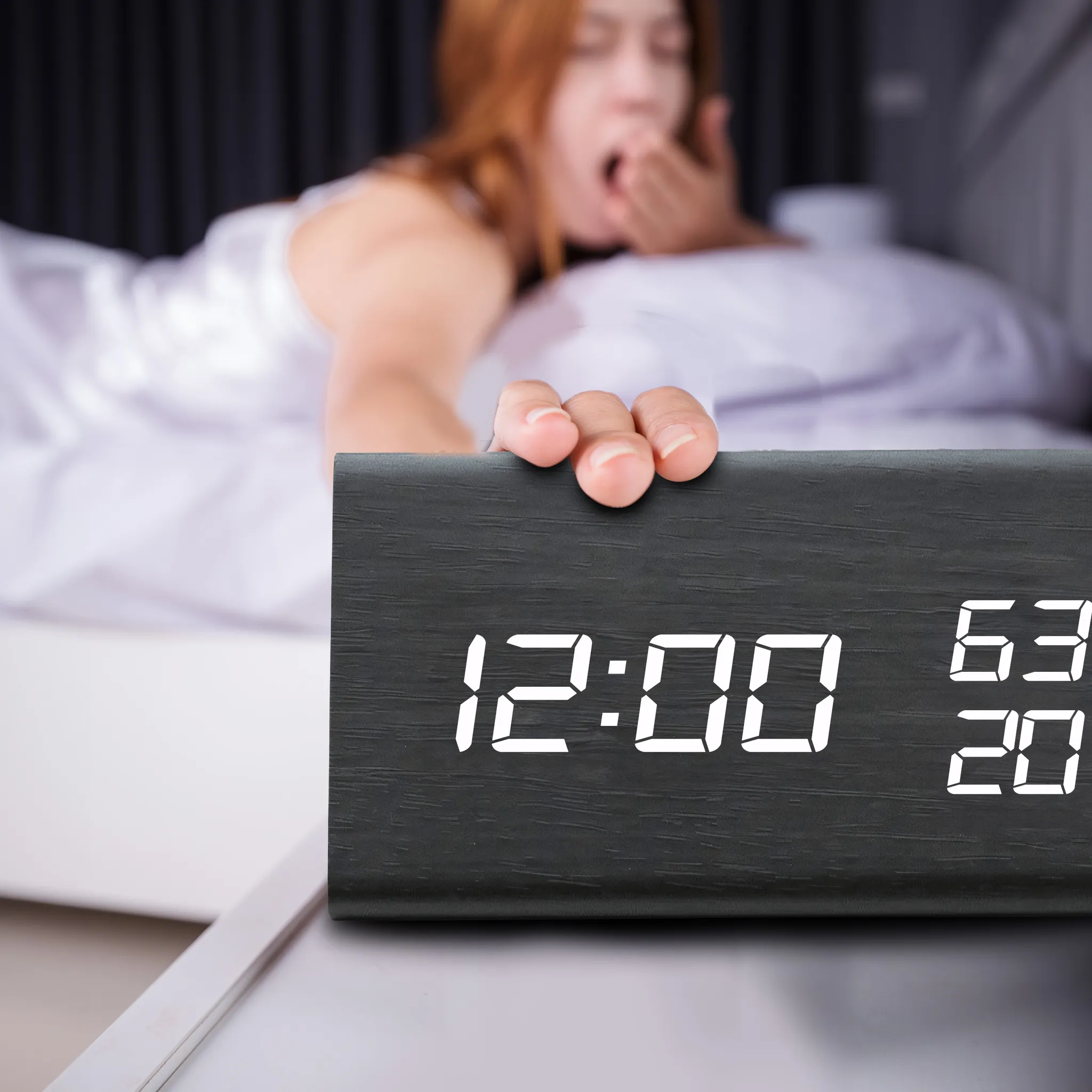 Eine Person gähnt und greift nach einer Digitaluhr mit der Anzeige 12:00. Die Uhr steht auf einem Nachttisch.