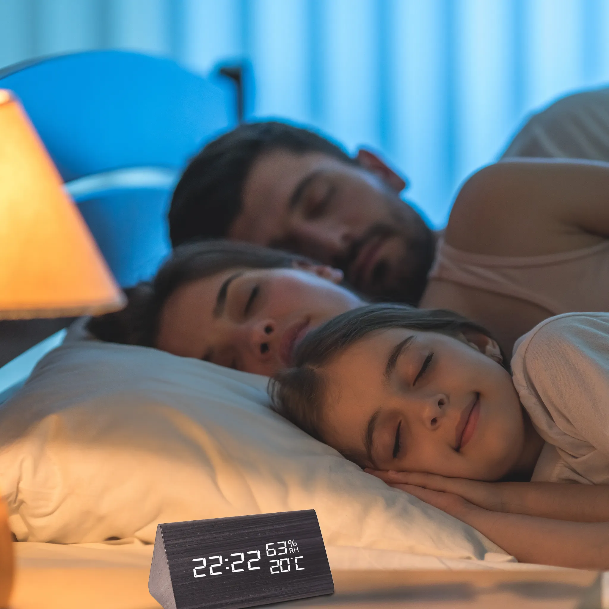 Familie schläft im Bett. Eine Digitaluhr zeigt die Zeit an. Eine Lampe ist an. Der Raum ist schwach beleuchtet.