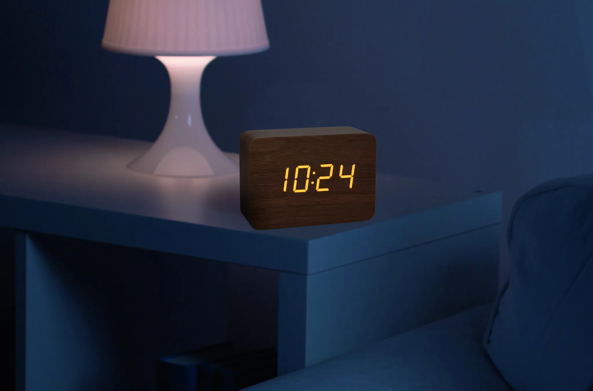 Eine Holzuhr zeigt 10:24. Eine weiße Lampe steht auf einem Tisch. Auf der rechten Seite ist ein weißes Sofa.