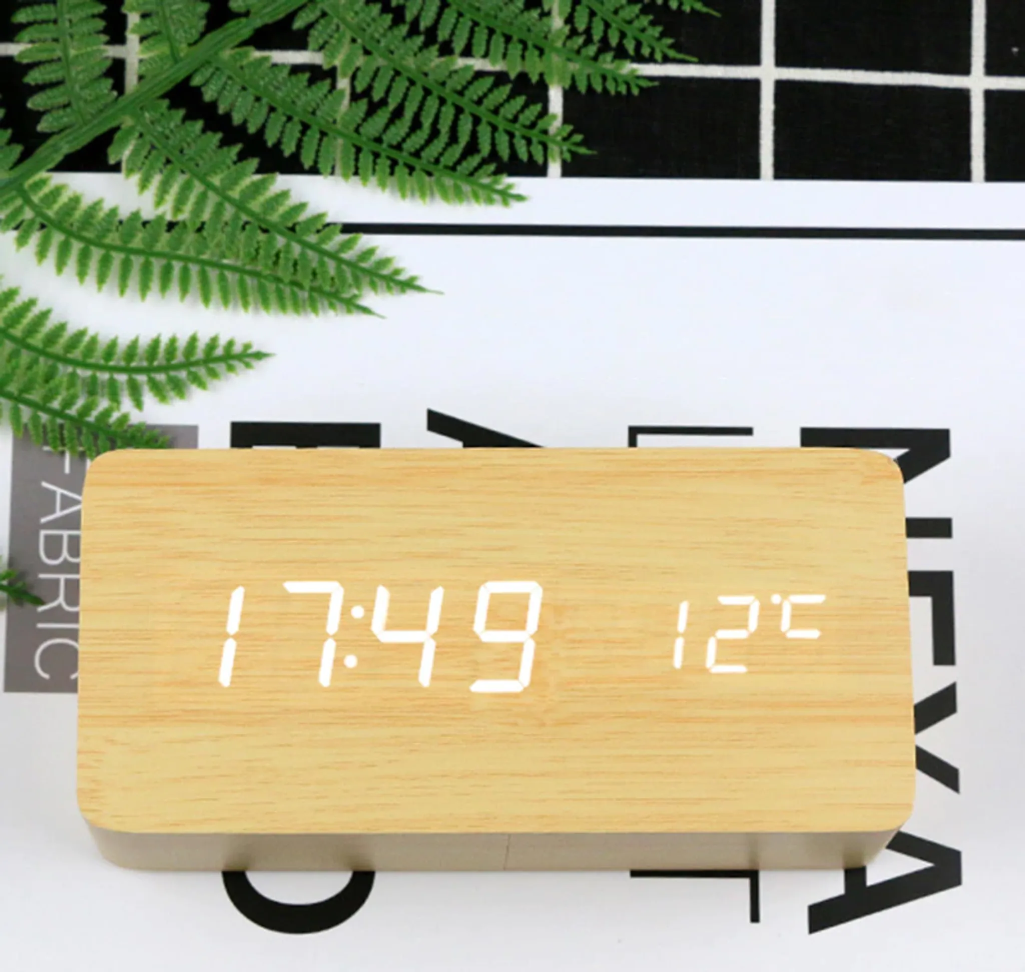 Eine digitale Holzuhr zeigt 17:49 und 12°C an. Oben links sind grüne Blätter, dahinter ein schwarz-weißer Hintergrund.