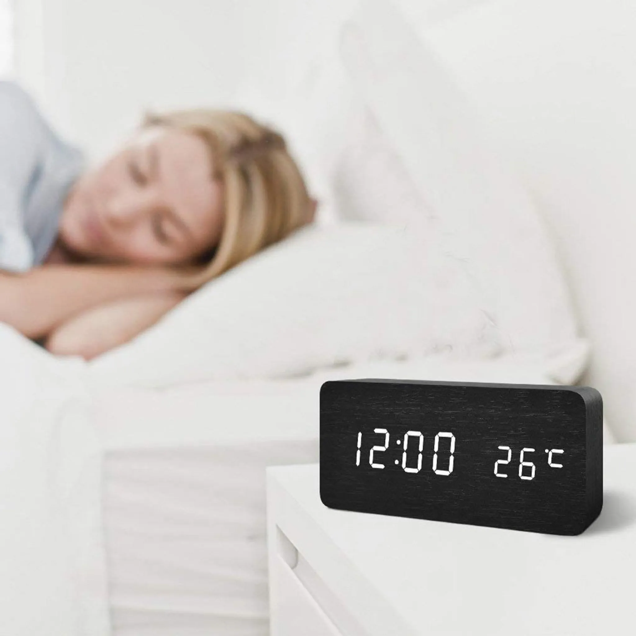 Eine Frau schläft im Bett. Eine schwarze Digitaluhr zeigt 12:00 und 26°C auf einem weißen Nachttisch an.