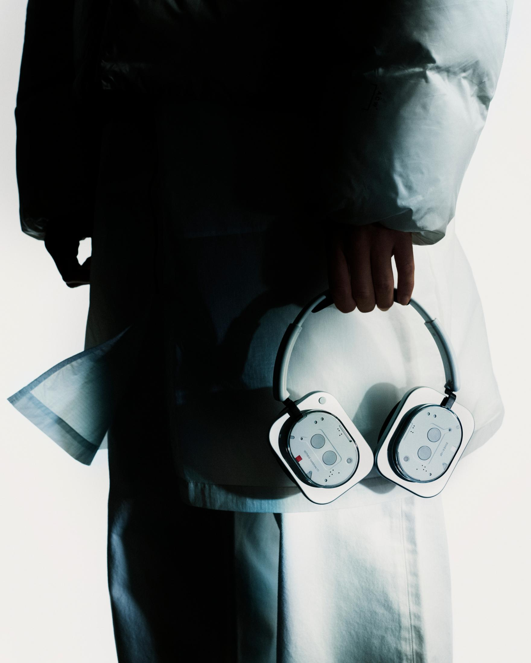 Osoba w bieli trzyma białe słuchawki. Tło jest białe, a osoba jest w cieniu.