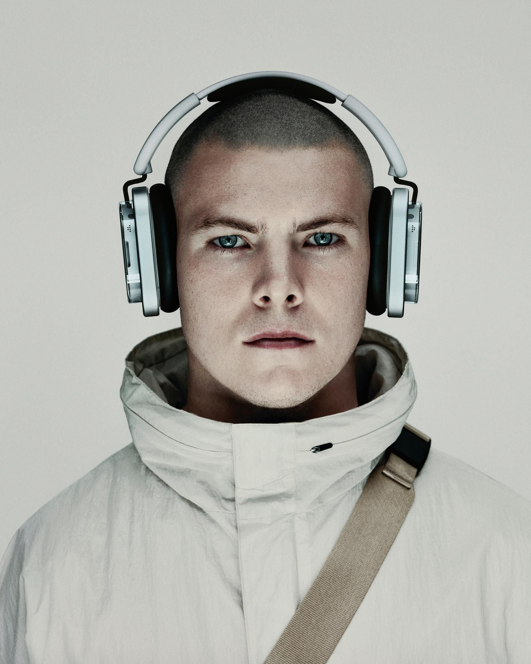 Mężczyzna z ogoloną głową w słuchawkach i jasnej kurtce, patrzący prosto w kamerę. Neutralne tło.