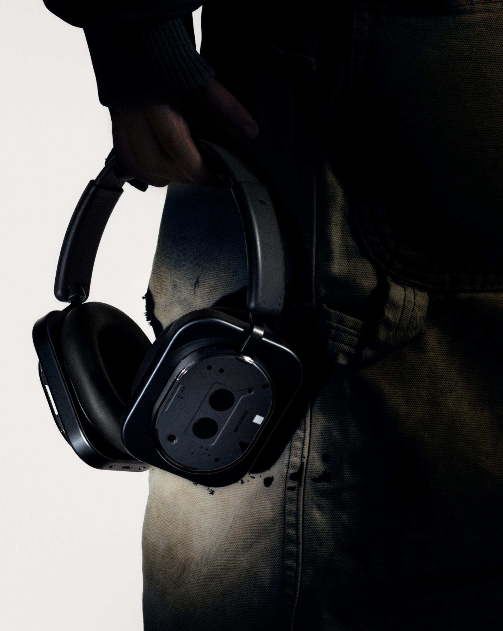 Osoba trzyma czarne słuchawki. Osoba ma na sobie zielony rękaw i zużyty, ciemny kombinezon. Białe tło.