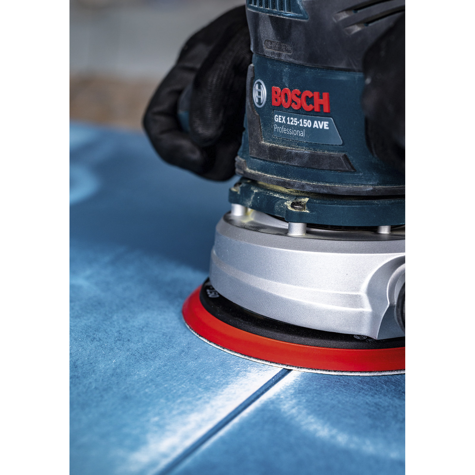 Ein Bosch-Schwingschleifer wird auf einer blauen Oberfläche verwendet. Eine behandschuhte Hand hält das Werkzeug. Der Schleifer hat ein rotes Pad.