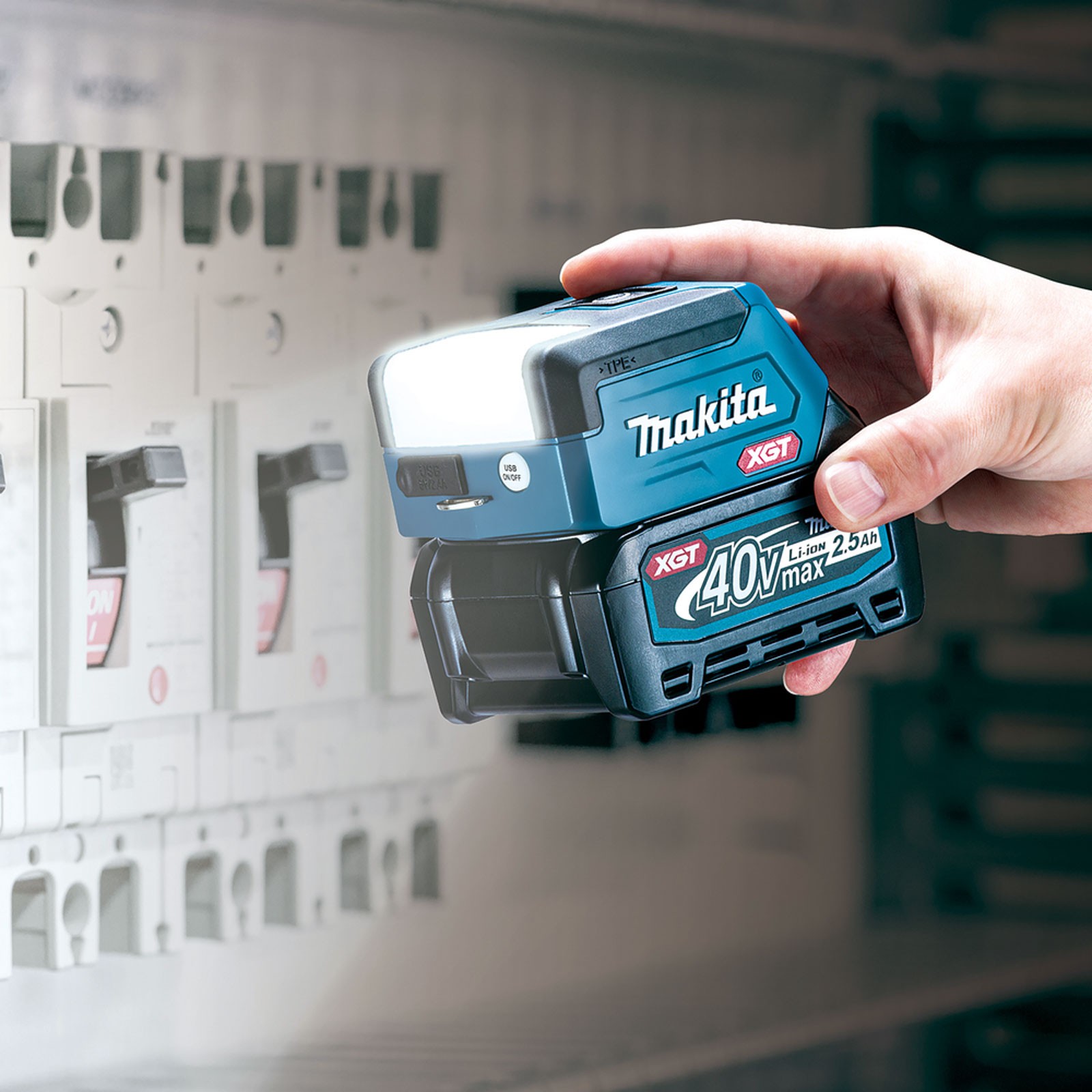 Ręka trzyma lampę roboczą Makita. Ma światło na górze i akumulator na dole. W tle znajdują się elementy elektryczne.