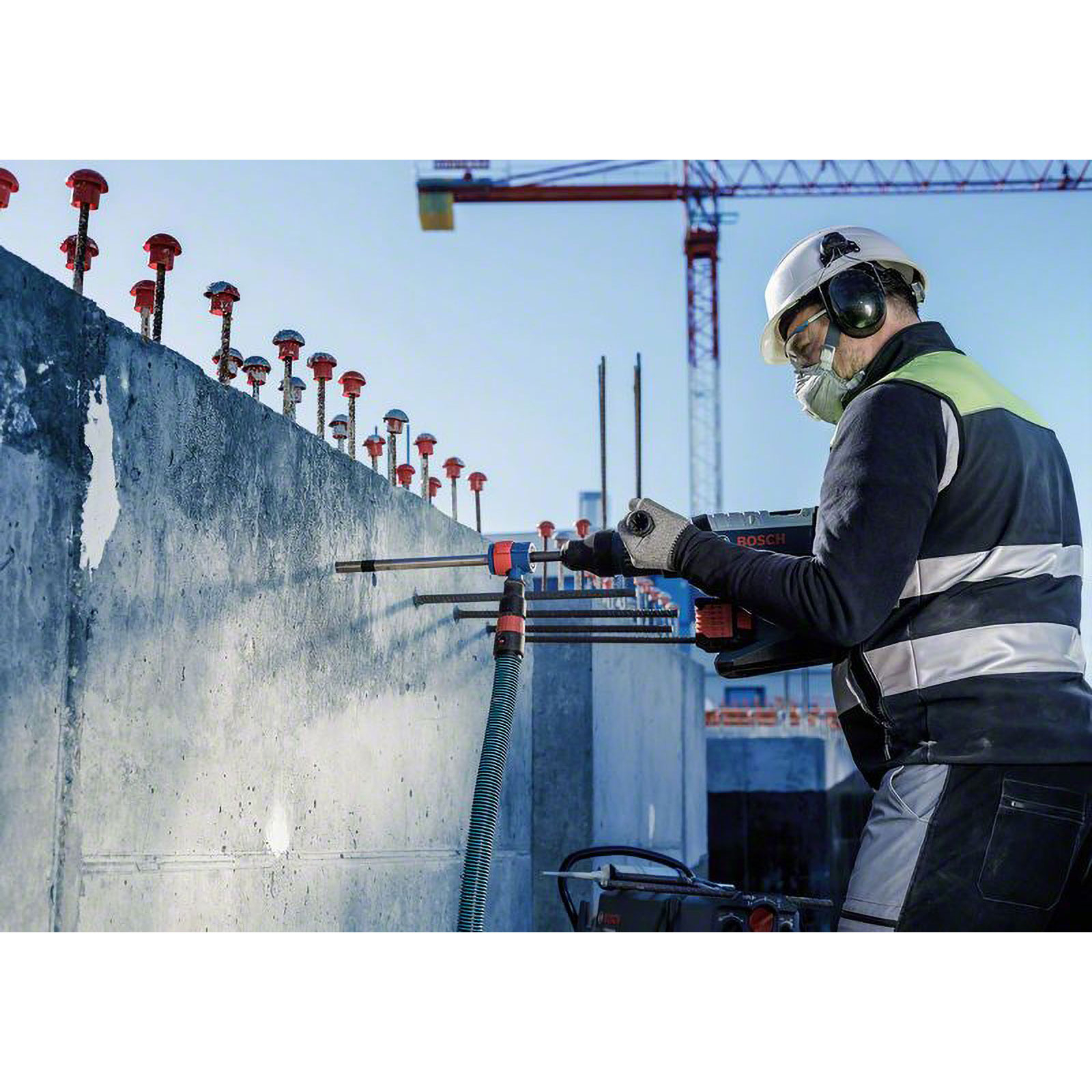 Un operaio edile fora un muro di cemento. Indossa casco, protezione per le orecchie e maschera. Una gru è sullo sfondo.