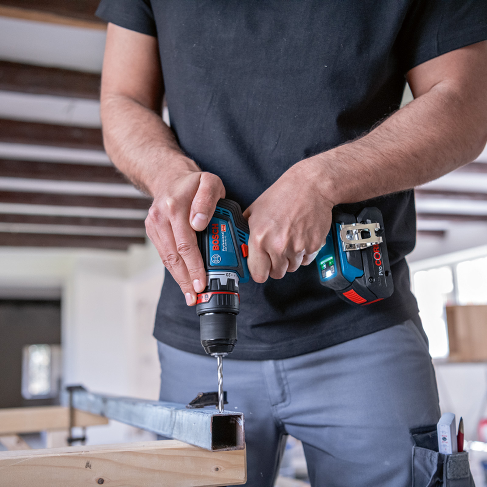 Osoba wierci w metalu niebiesko-czarną wiertarką Bosch. Ma na sobie czarną koszulę i szare spodnie. W tle drewniana konstrukcja.