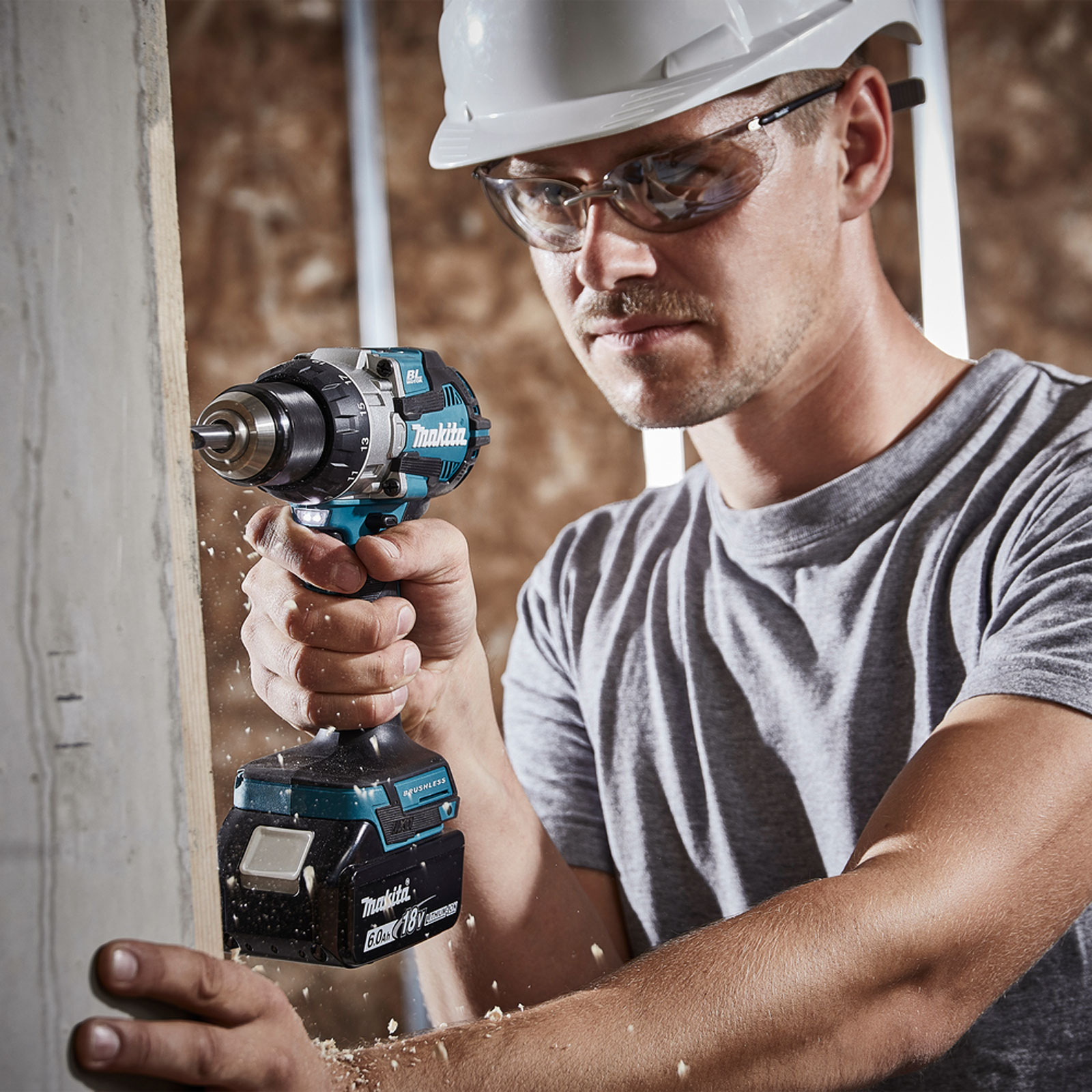 Ein Bauarbeiter bohrt mit einem Makita-Bohrer in Holz. Er trägt eine Schutzbrille und einen Helm. Sägemehl ist sichtbar.