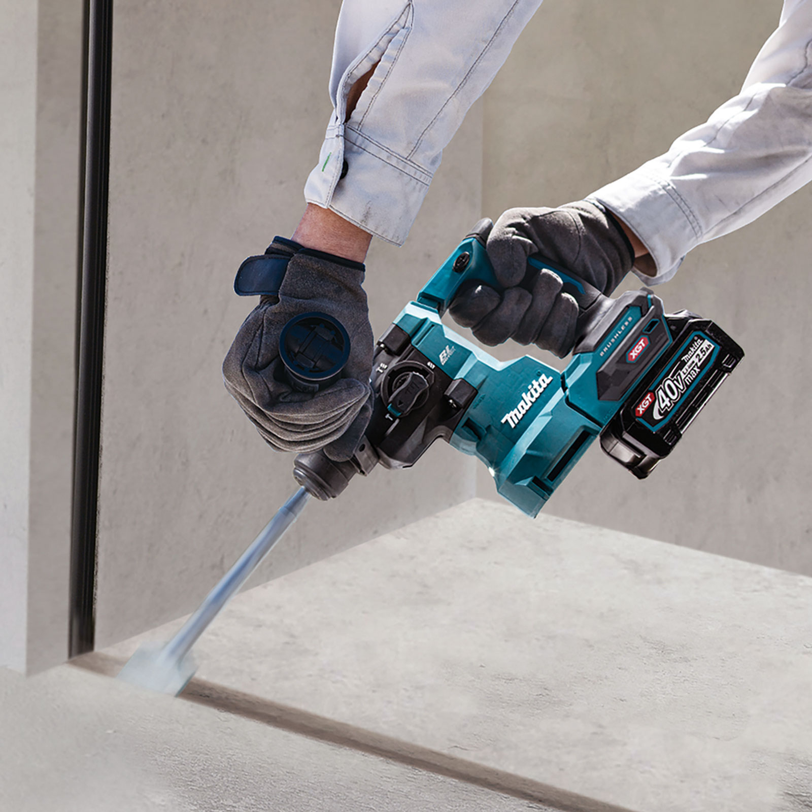 Ein Arbeiter benutzt einen Makita-Hammerbohrer in Teal und Schwarz auf Beton. Graue Handschuhe und hellblaue Hemdsärmel sind sichtbar.