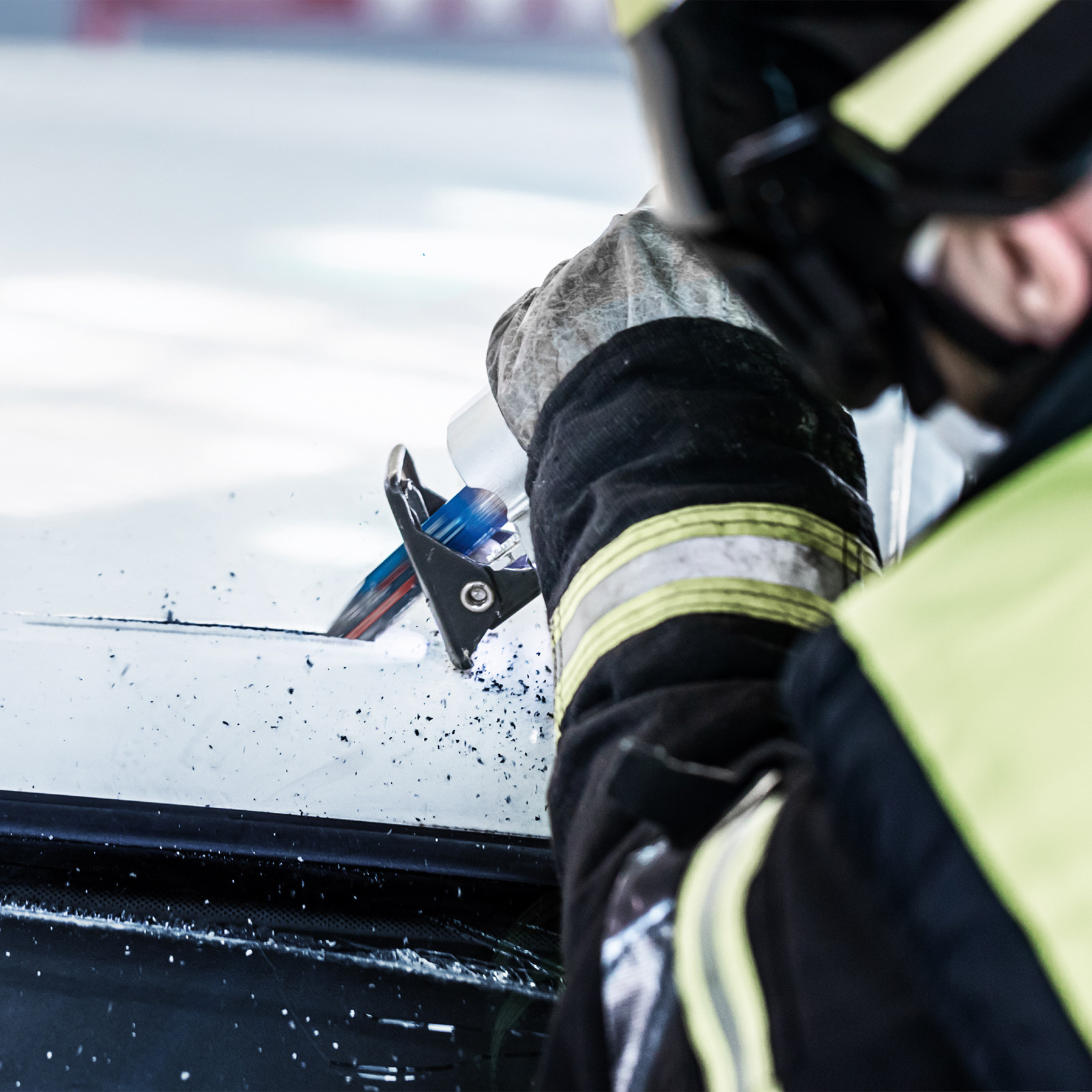 Strażak przecina szybę samochodową narzędziem. Scena pokazuje akcję z bliska, z niebiesko-czerwonym paskiem.