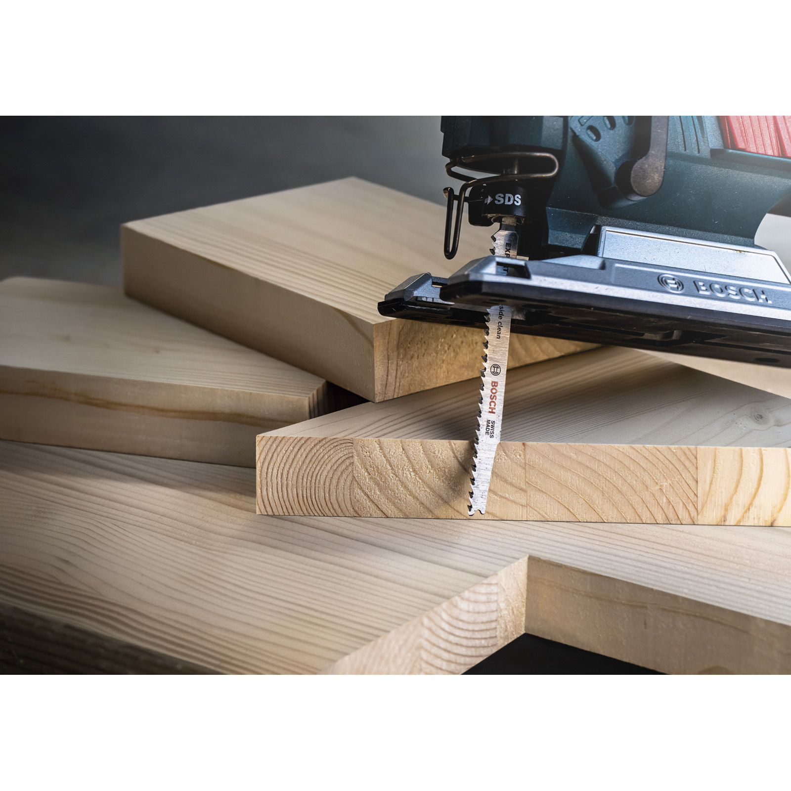 Wyrzynarka tnie drewnianą deskę. Narzędzie jest ciemnozielone i czarne. Kilka drewnianych desek w tle.