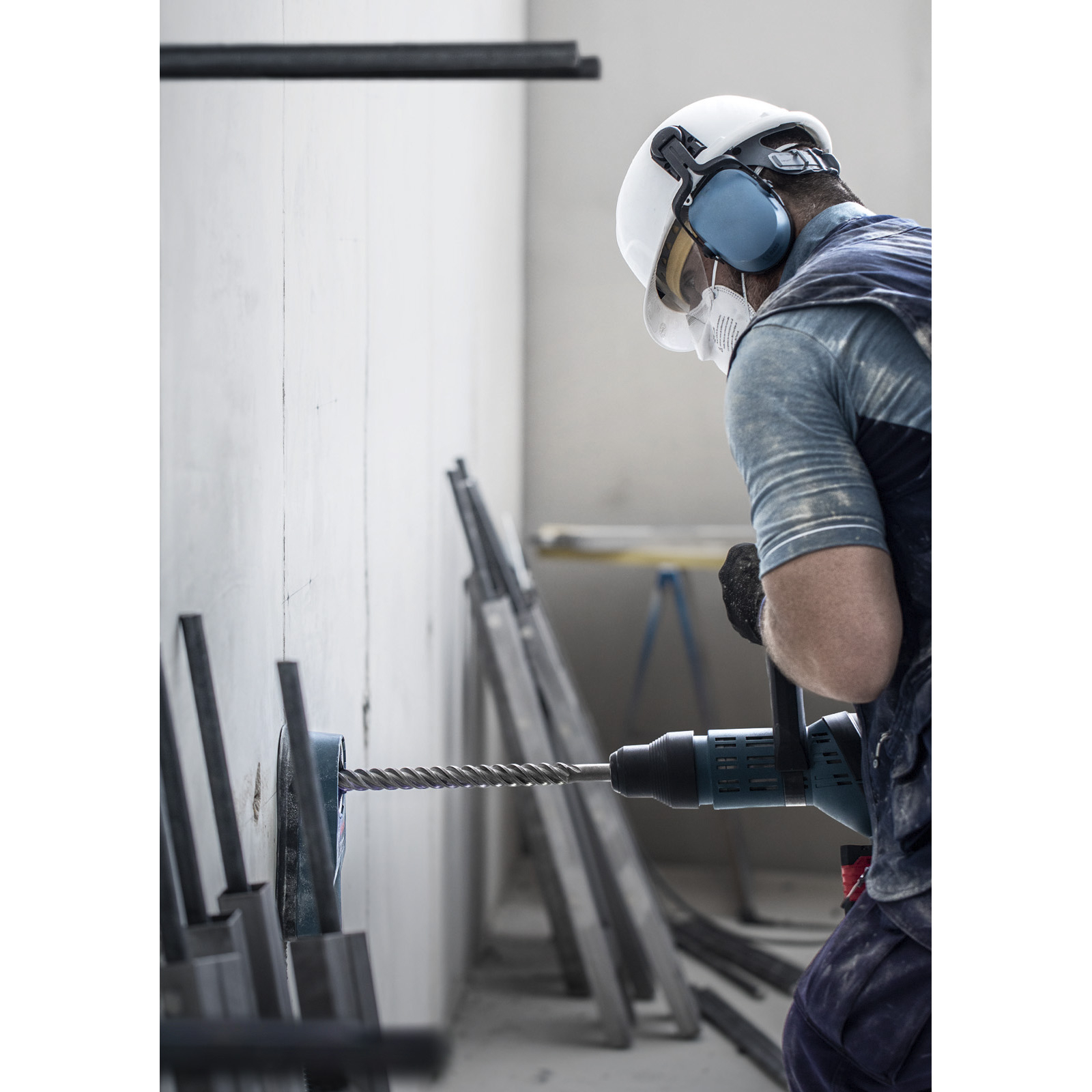 Robotnik wierci w betonowej ścianie wiertarką. Ma na sobie kask, ochronniki słuchu i maskę.