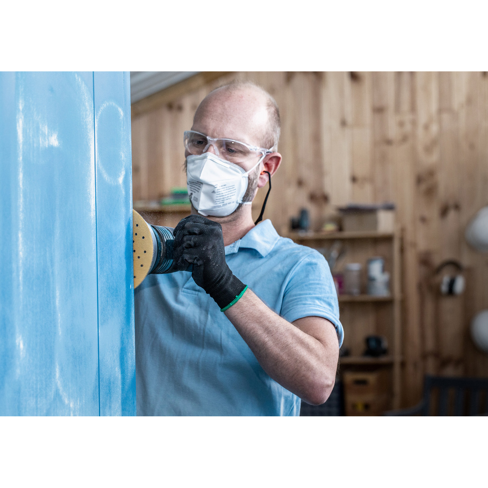 Ein Mann schleift eine blaue Oberfläche, trägt Schutzbrille, Maske und Handschuhe in einer Werkstatt.