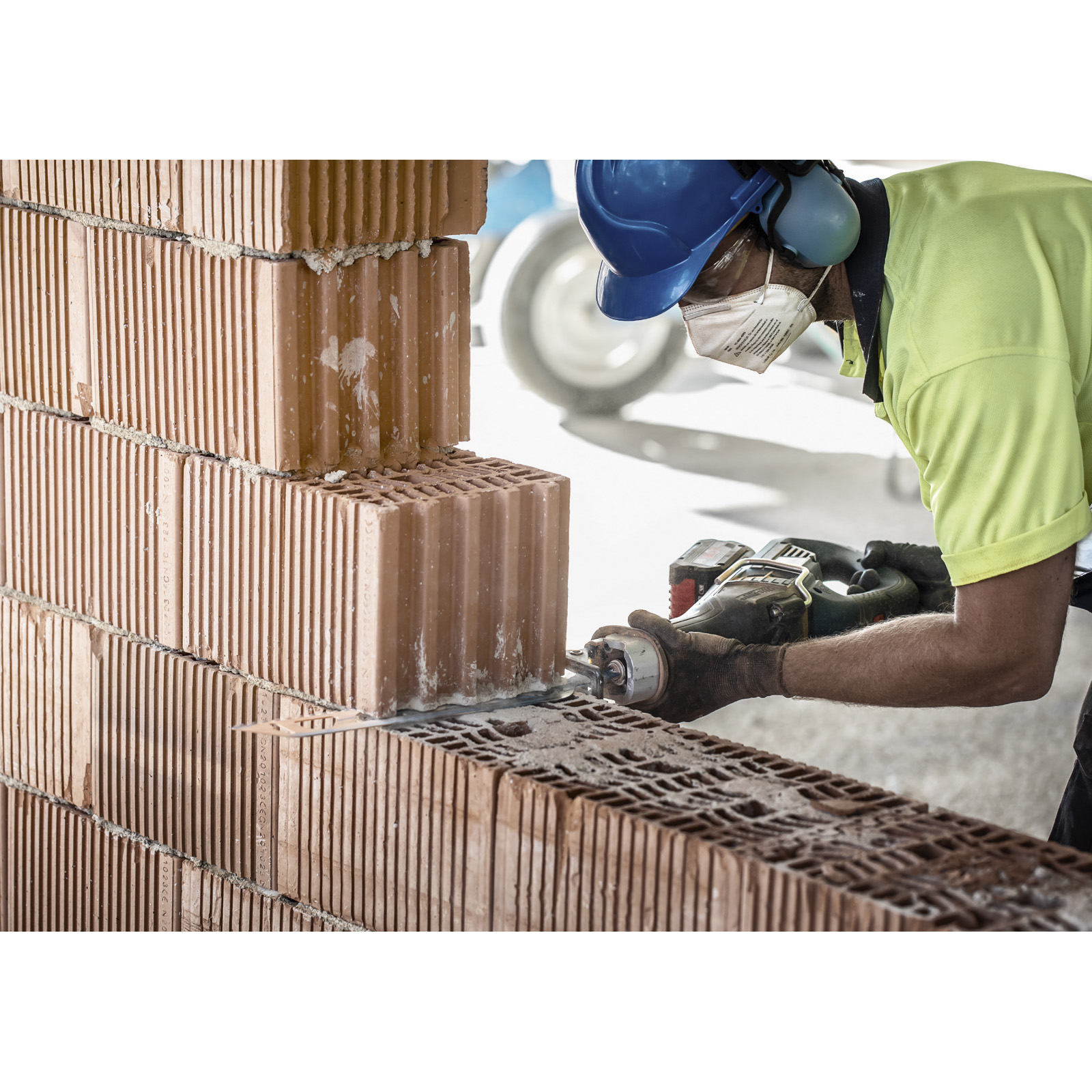 Robotnik budowlany tnie ceglaną ścianę piłą. Pracownik nosi kask, ochronniki słuchu i maskę.