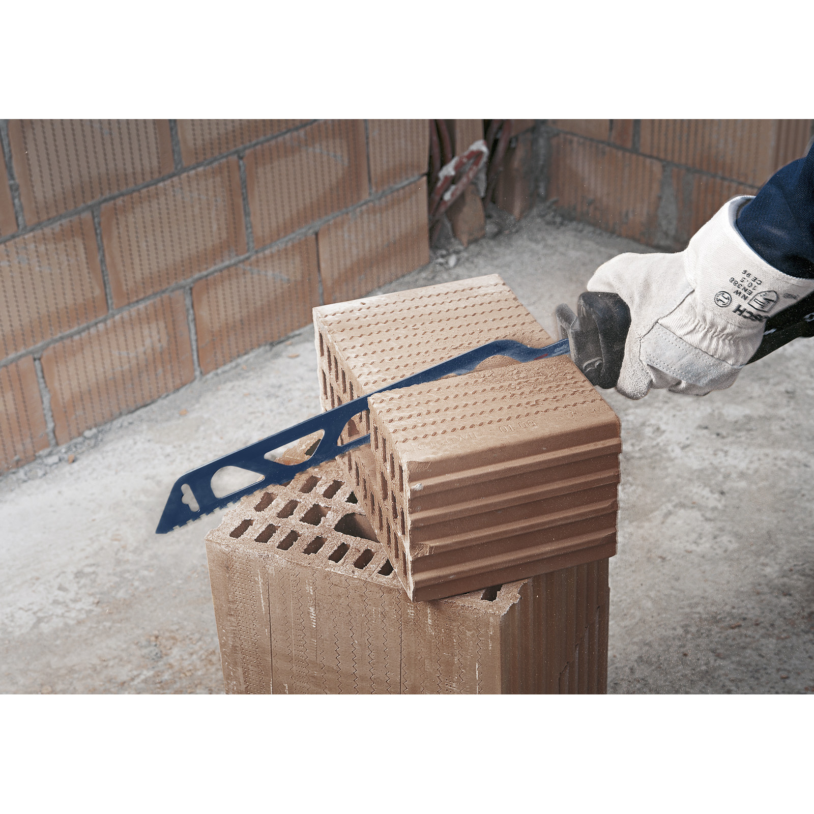 Osoba piłuje cegłę niebieską piłą. Cegła jest na innej cegle. Tłem jest ceglany mur.