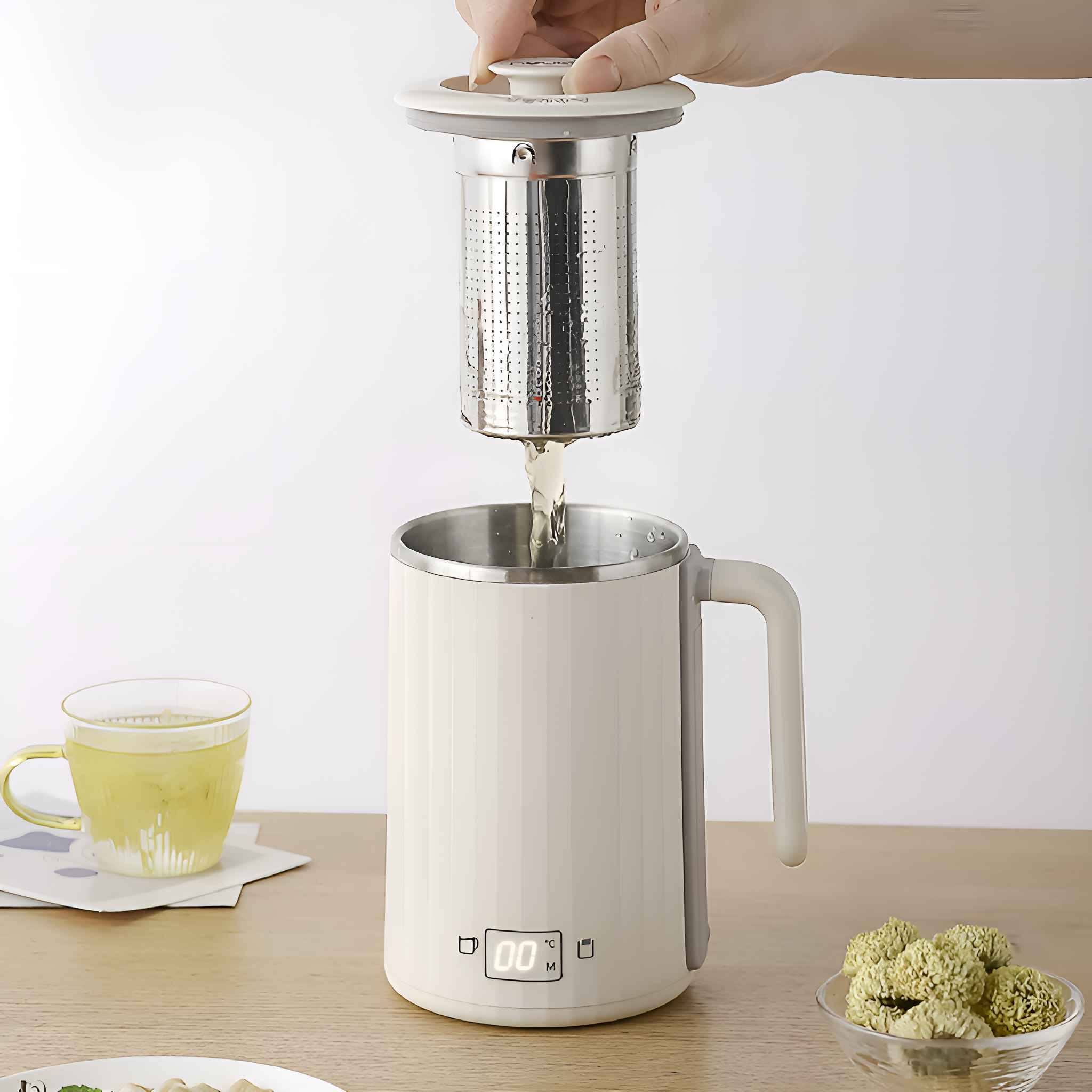 Eine Hand gießt Flüssigkeit aus einem Tee-Edelstahl-Sieb in einen weißen Becher. Eine Tasse Tee, eine Schüssel mit Kräutern und Essen sind ebenfalls vorhanden.
