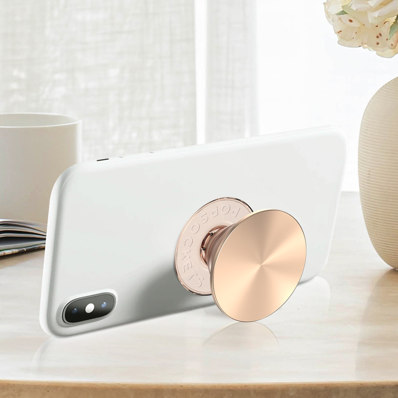 Weißes Telefon mit roségoldenem PopSocket auf einem Tisch. Im Hintergrund eine Tasse und eine Vase mit Blumen.