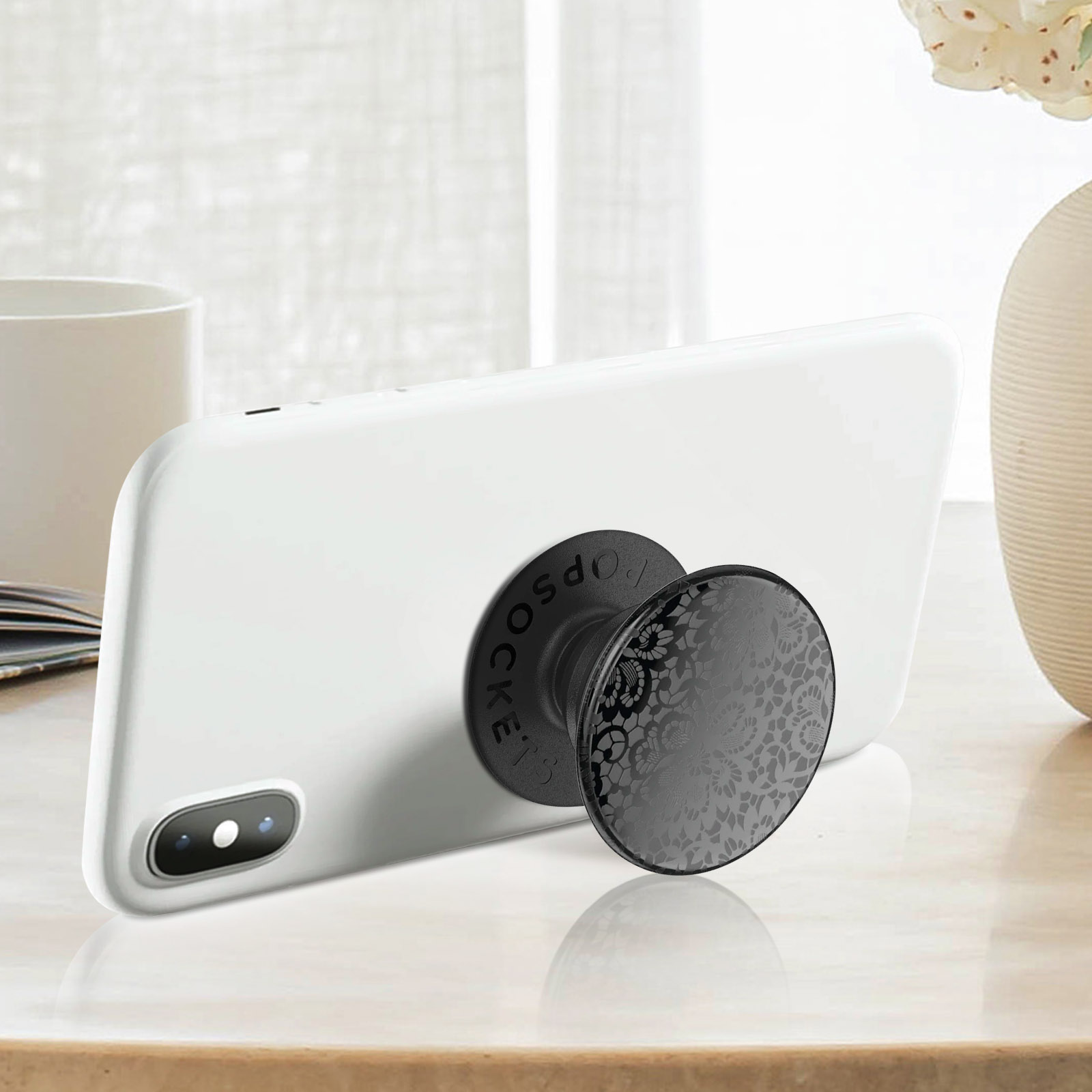 Un telefono bianco con un PopSocket nero con un design floreale è su un tavolo. Un vaso e una tazza sono sullo sfondo.