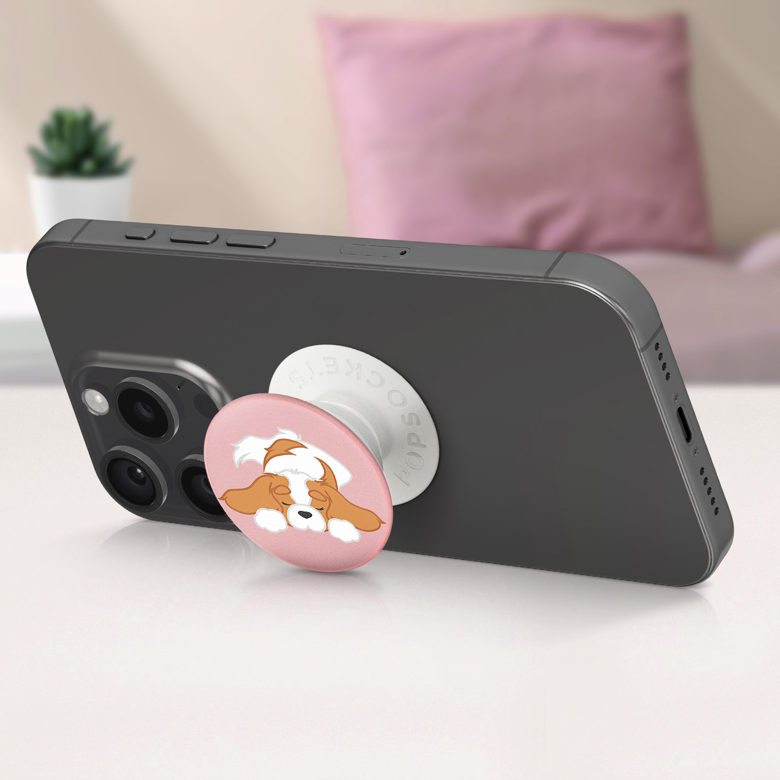 Un telefono nero con un PopSocket che mostra un cane dei cartoni animati. Lo sfondo ha una pianta, un letto e un cuscino rosa.