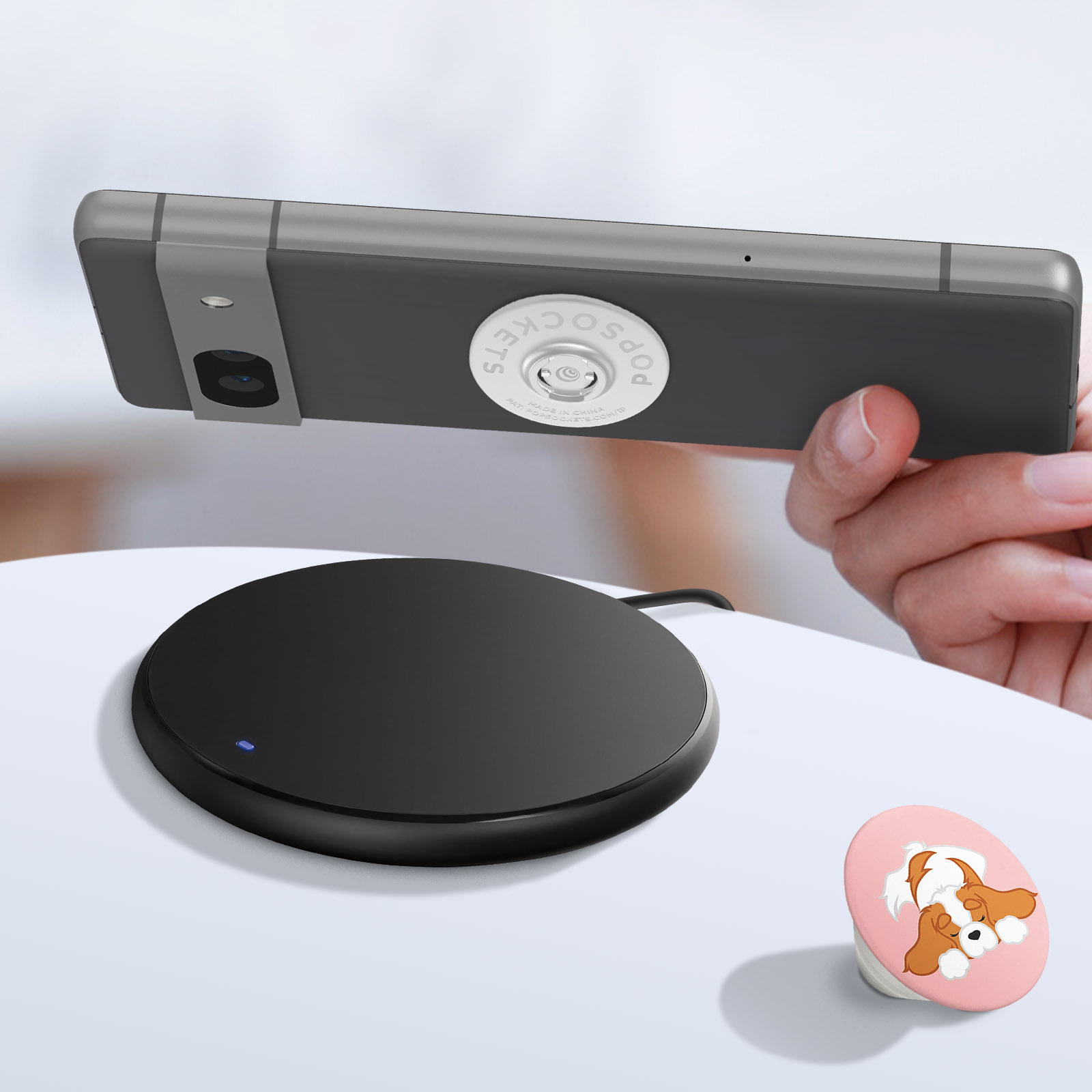 Un telefono con PopSocket è tenuto sopra un caricatore wireless nero. Un PopSocket a forma di cane è su una superficie bianca.